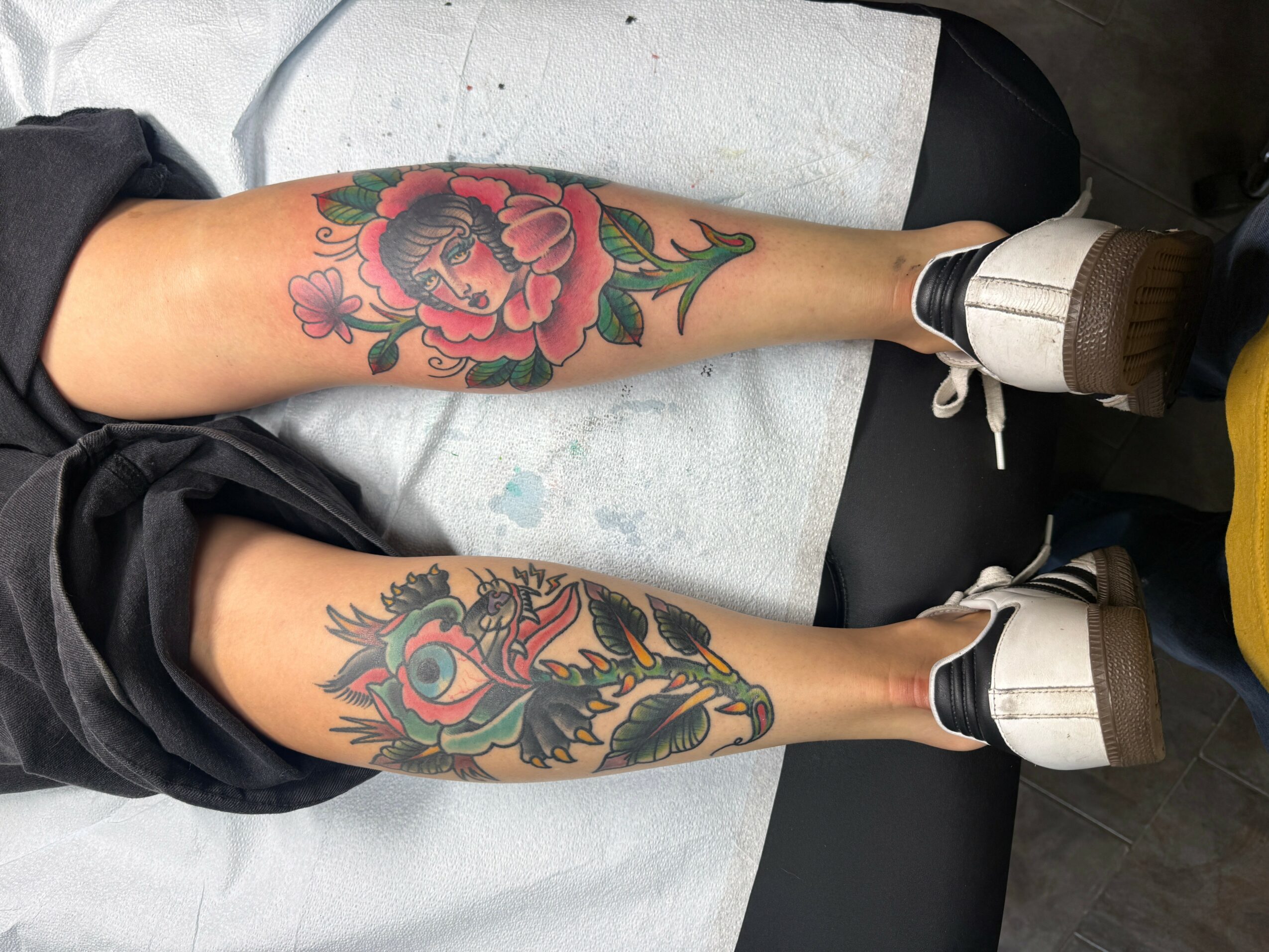 Two legs with colorful tattoos, one with a pink flower and green leaves, the other with a dragon and eye design.