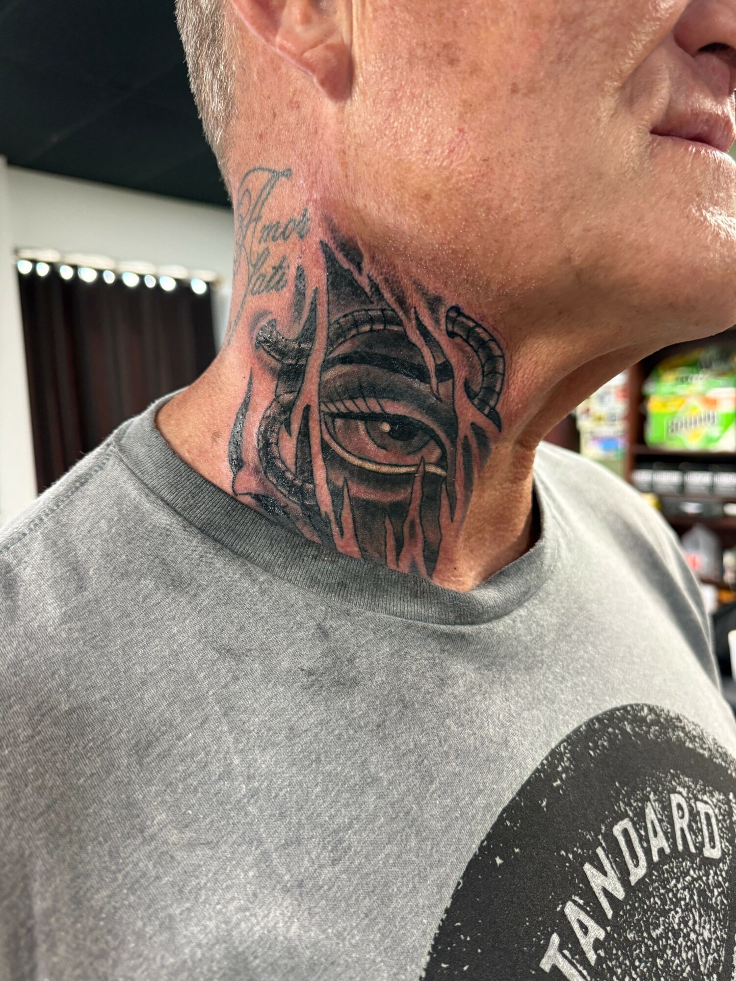 Person with a tattoo of an eye on their neck, wearing a gray shirt, in an indoor setting.