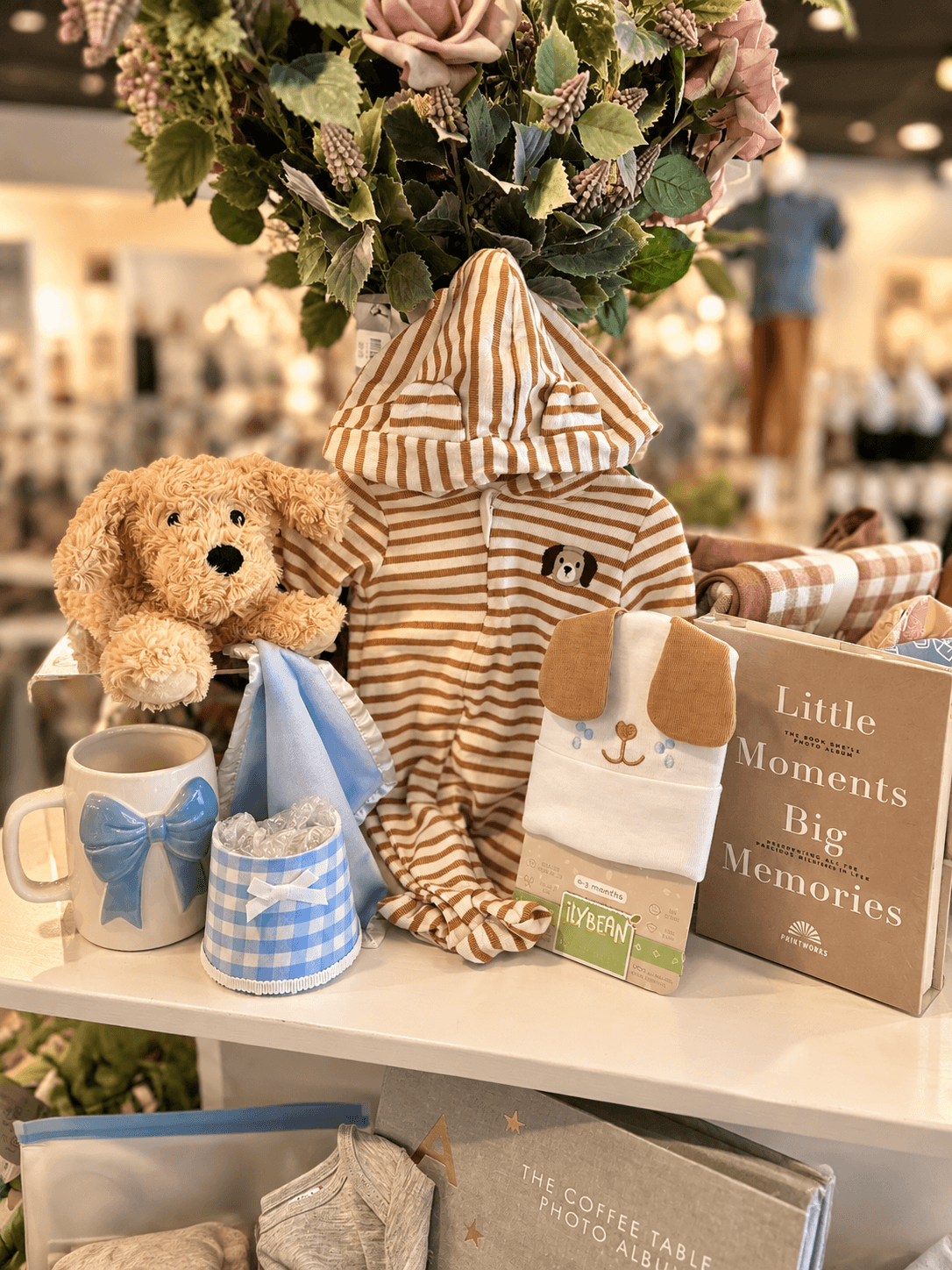Striped baby outfit, teddy bear, baby shoes, blue accessories, and a book displayed on a shelf beneath flowers.