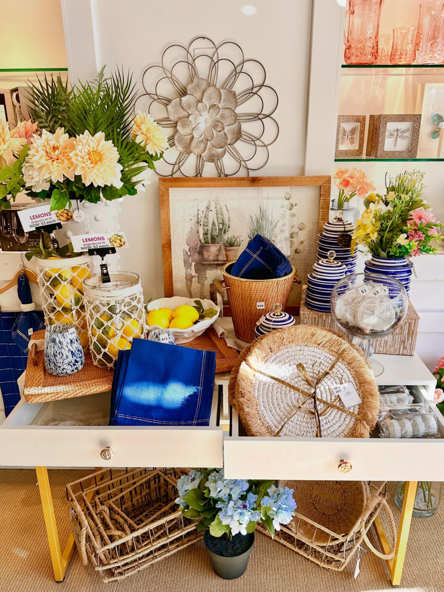 Decorative home goods display with flowers, framed art, stacked plates, baskets, and a metal wall sculpture.