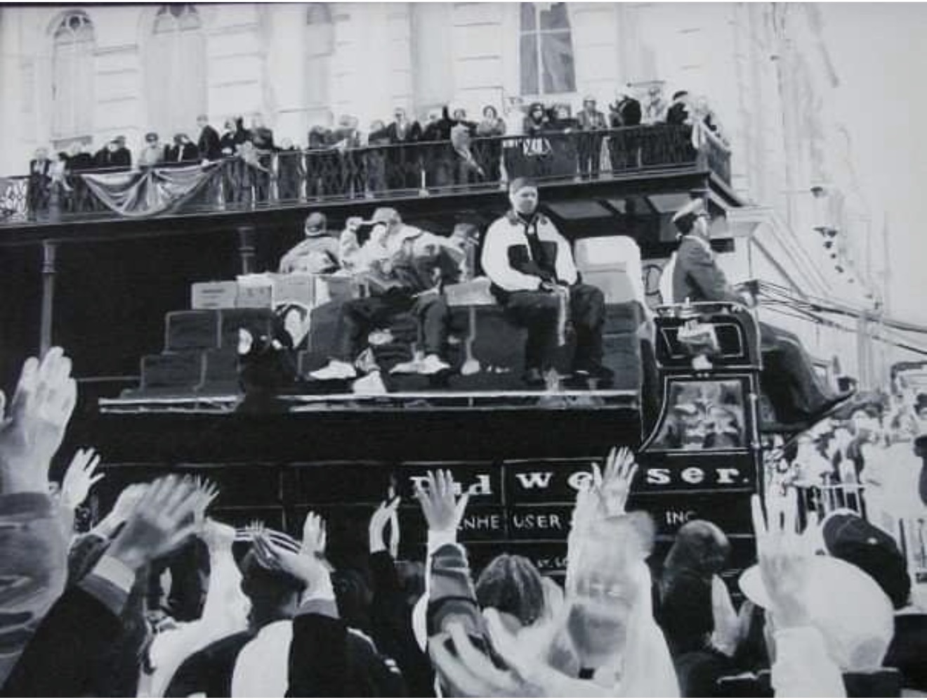 Black-and-white crowd reaches toward people seated on a vehicle, with spectators watching from an upper balcony.