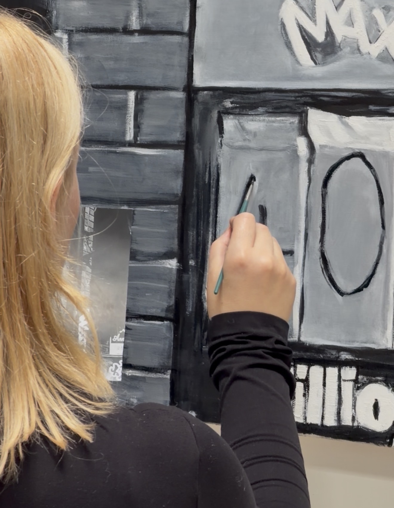 A person with long blond hair paints a black outline on a gray mural.