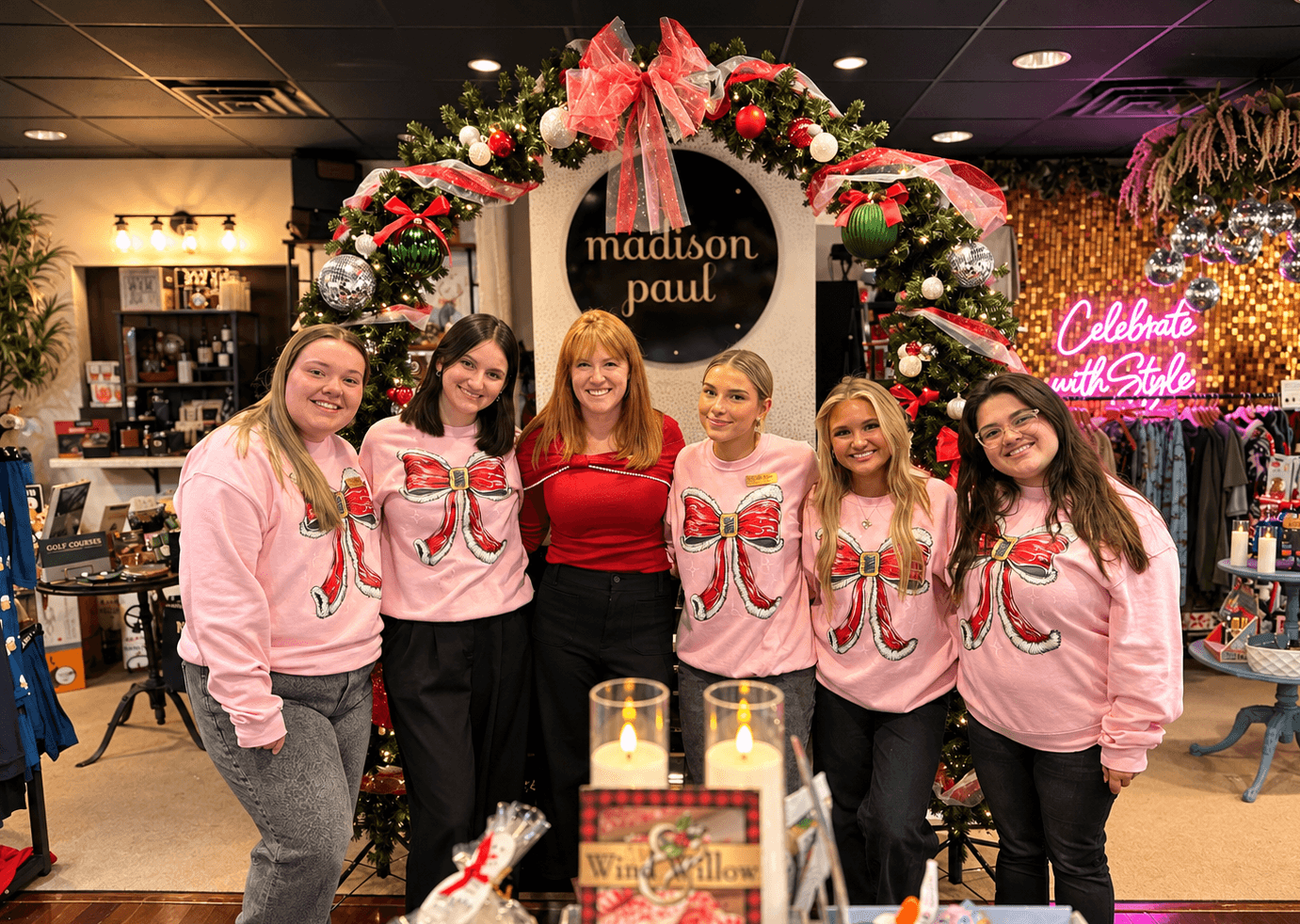 Six people pose in front of a decorated holiday arch with ornaments and a circular sign.
