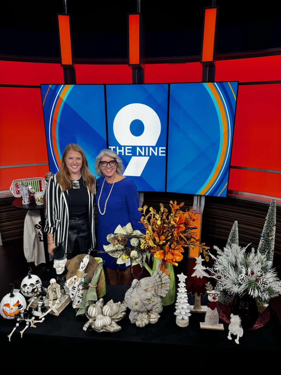 Two people stand behind a table of flowers and Halloween decorations in front of a blue The Nine backdrop.