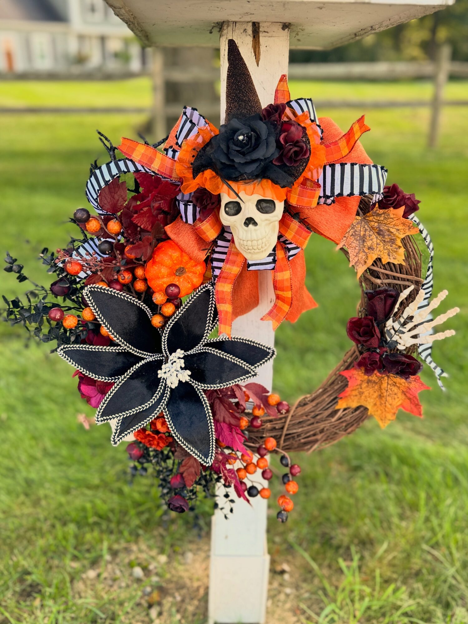 Decorative Halloween wreath with a skull, black flower, orange fabric, striped bows, leaves, and berries on a post.