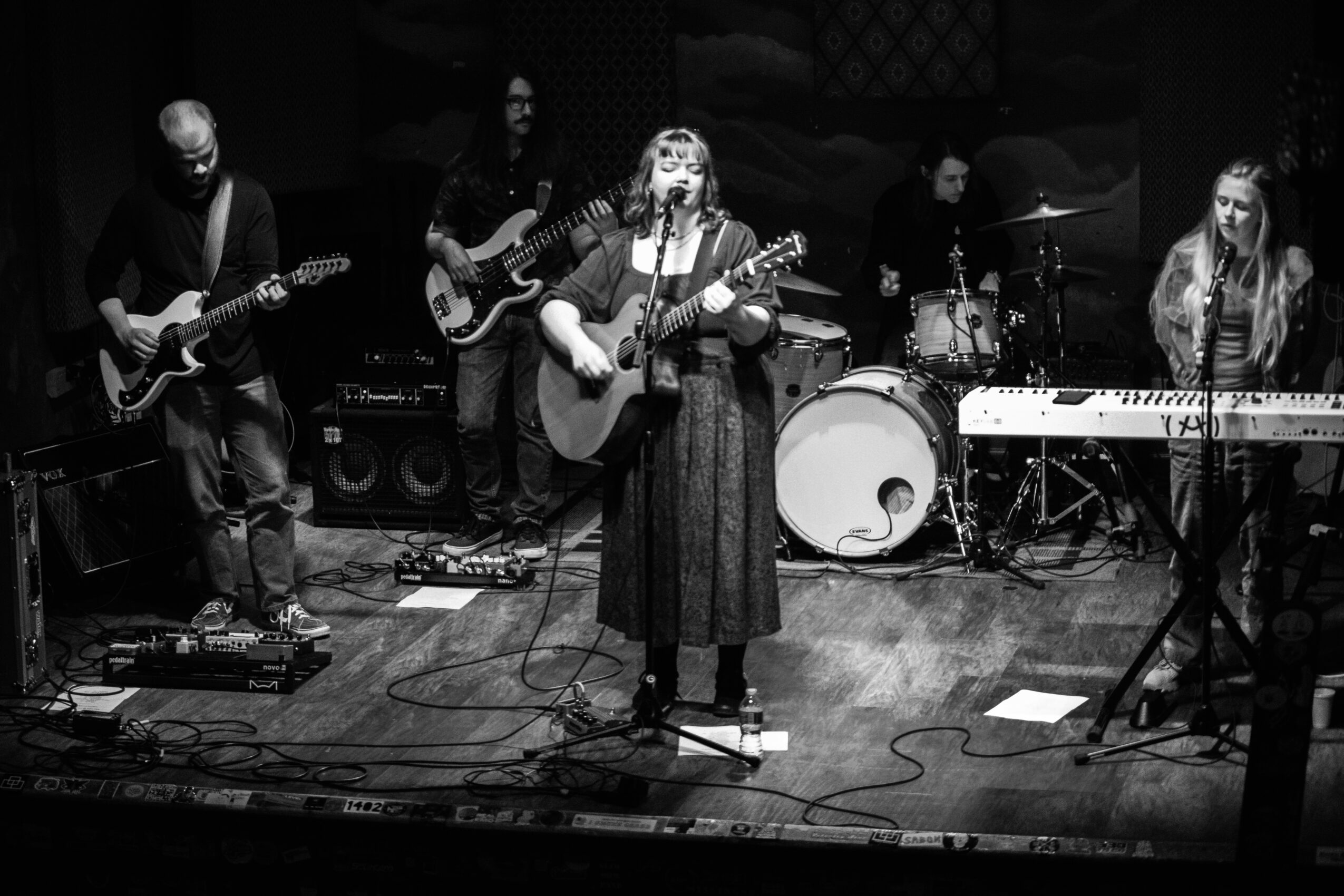 Group of five musicians performing on stage with instruments, including guitar, keyboard, and drums, in a black-and-white photo.