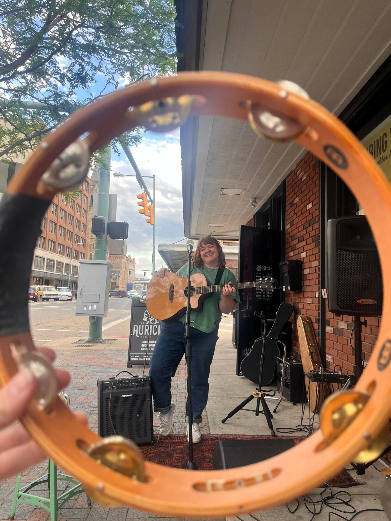 Person playing guitar on a small stage outdoors, with a brick building and trees in the background.