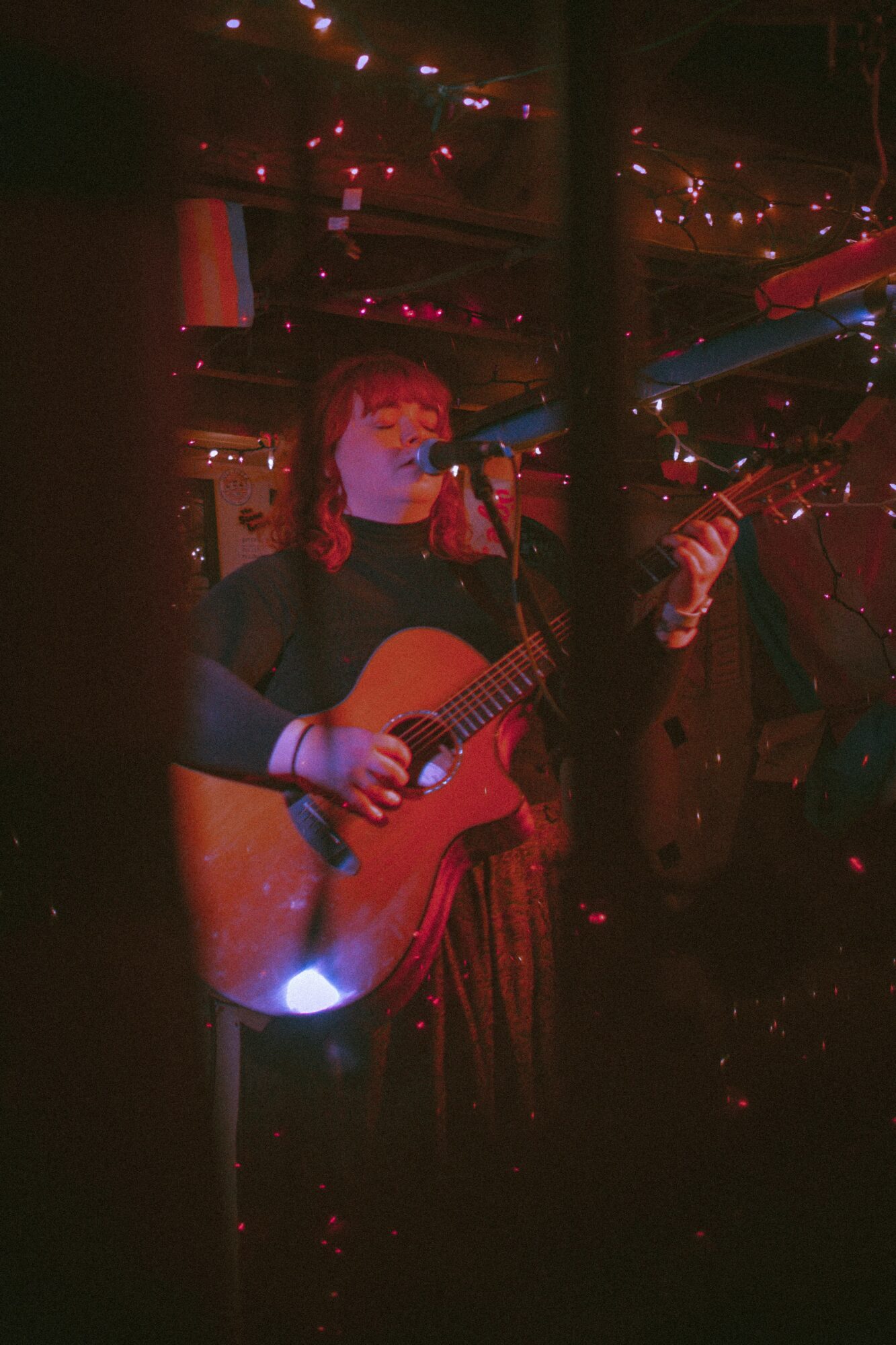 Person playing guitar and singing into microphone in dimly lit setting with string lights overhead.