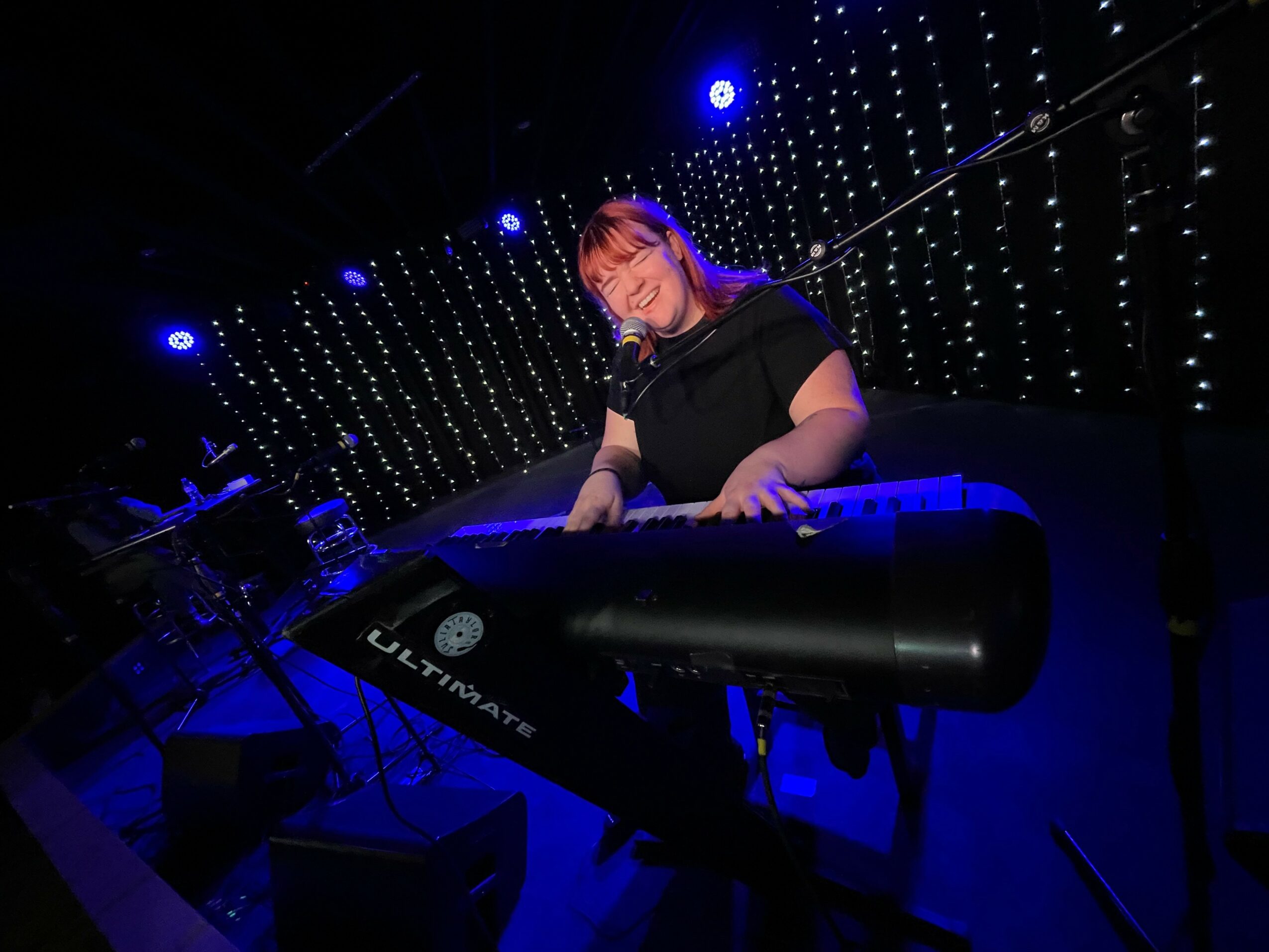 Person playing keyboard on stage with blue lighting and decorative background lights, smiling, wearing black shirt.