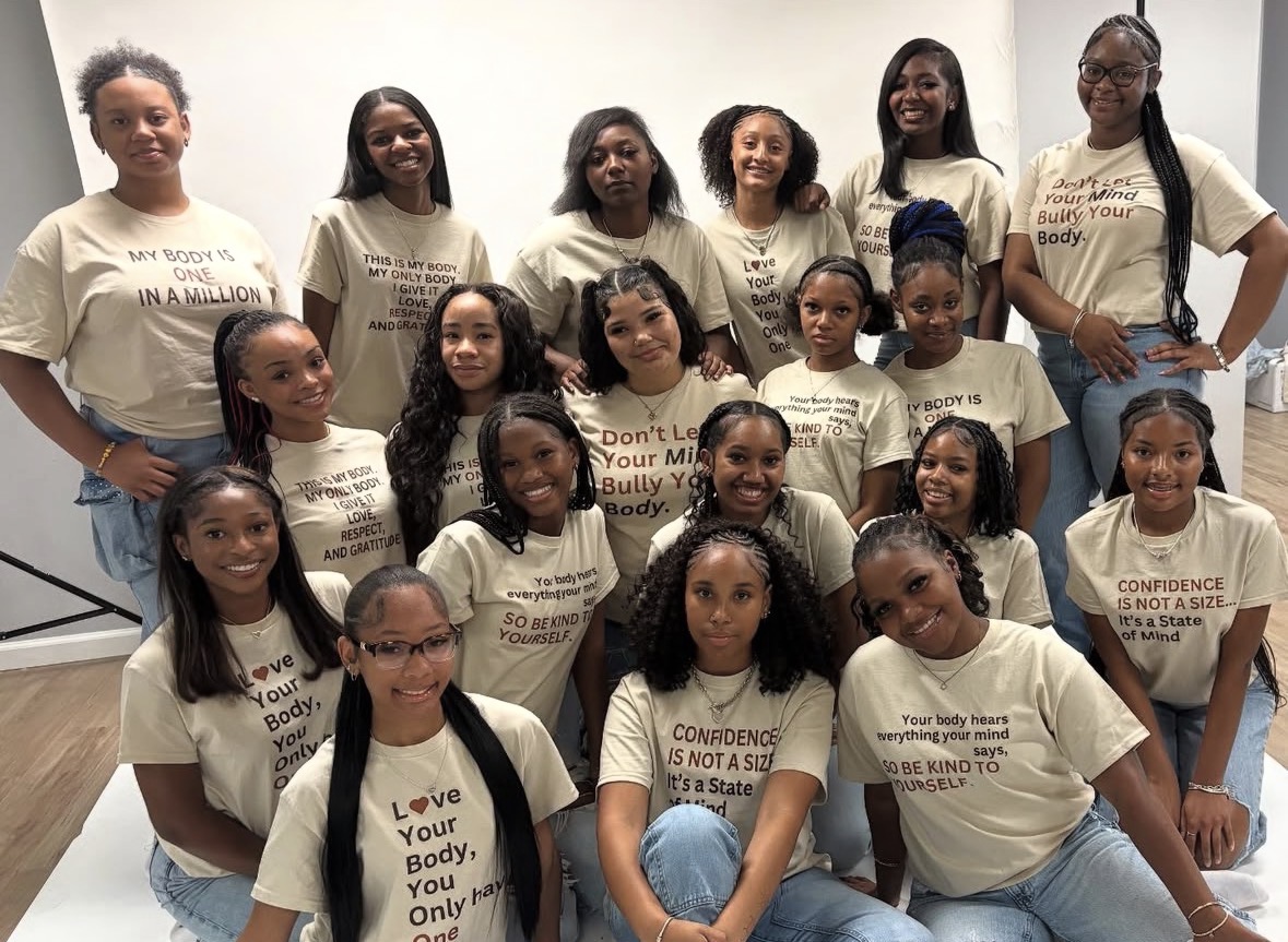 Group of 16 young girls standing and sitting together, wearing matching beige T-shirts, smiling, against a plain background.