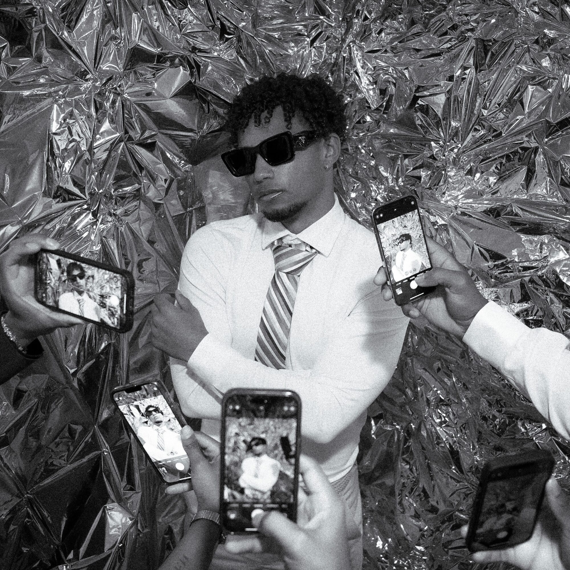 A person in sunglasses and a striped tie poses against crumpled reflective foil as others photograph them with phones.