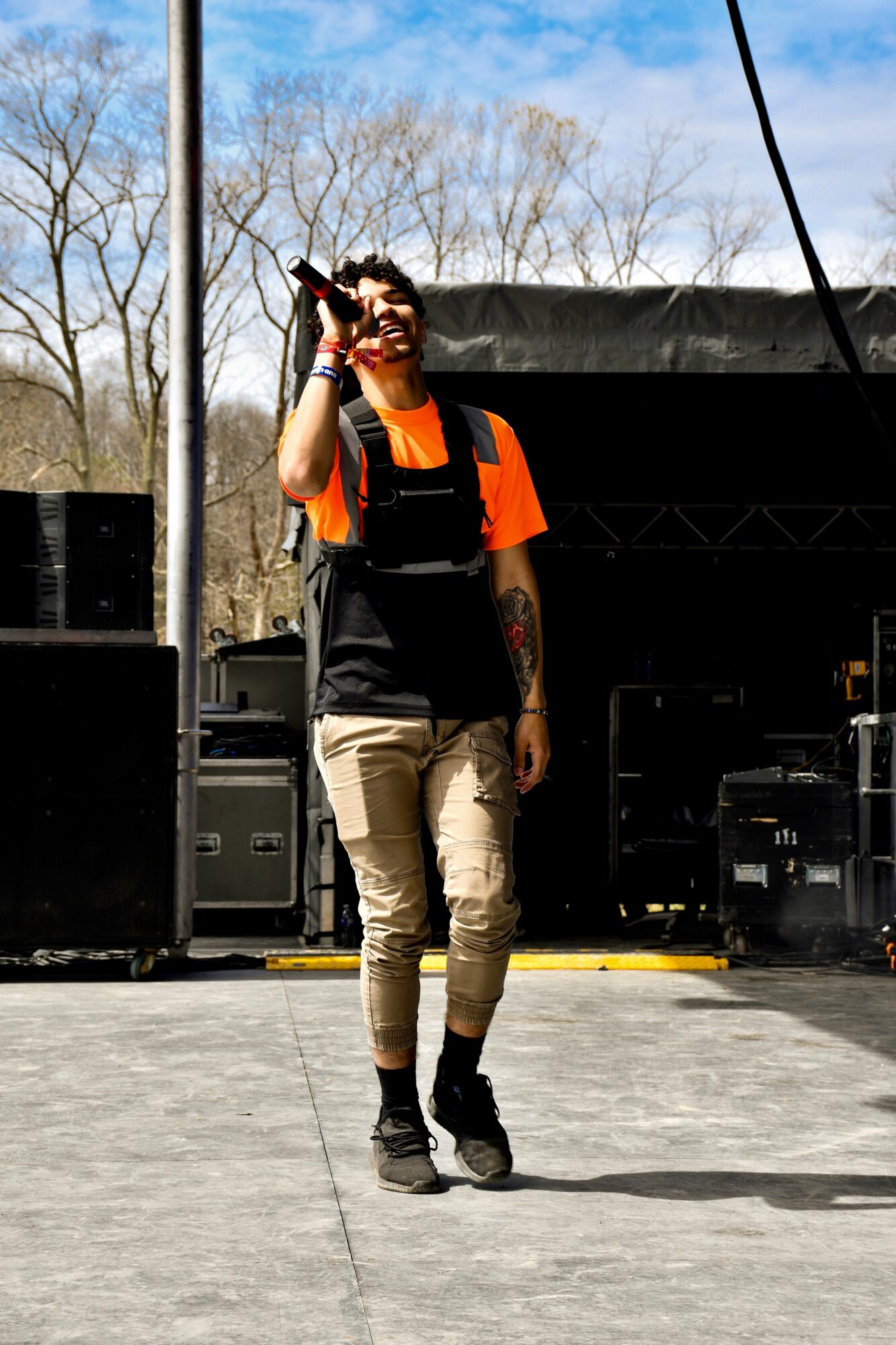 Person in an orange shirt and black vest holds a microphone on an outdoor stage.