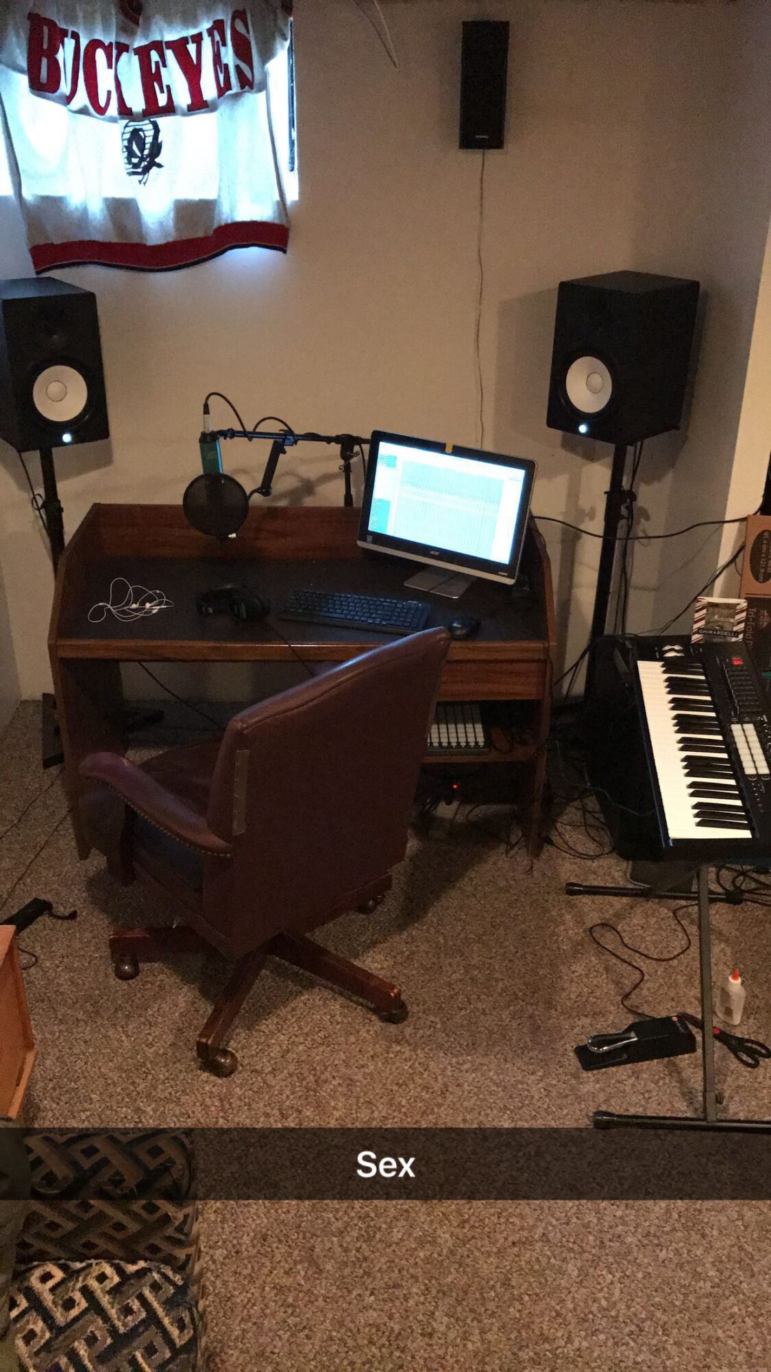 Small recording setup with a desk, computer monitor, speakers, microphone, chair, and keyboard; a banner hangs on the wall.