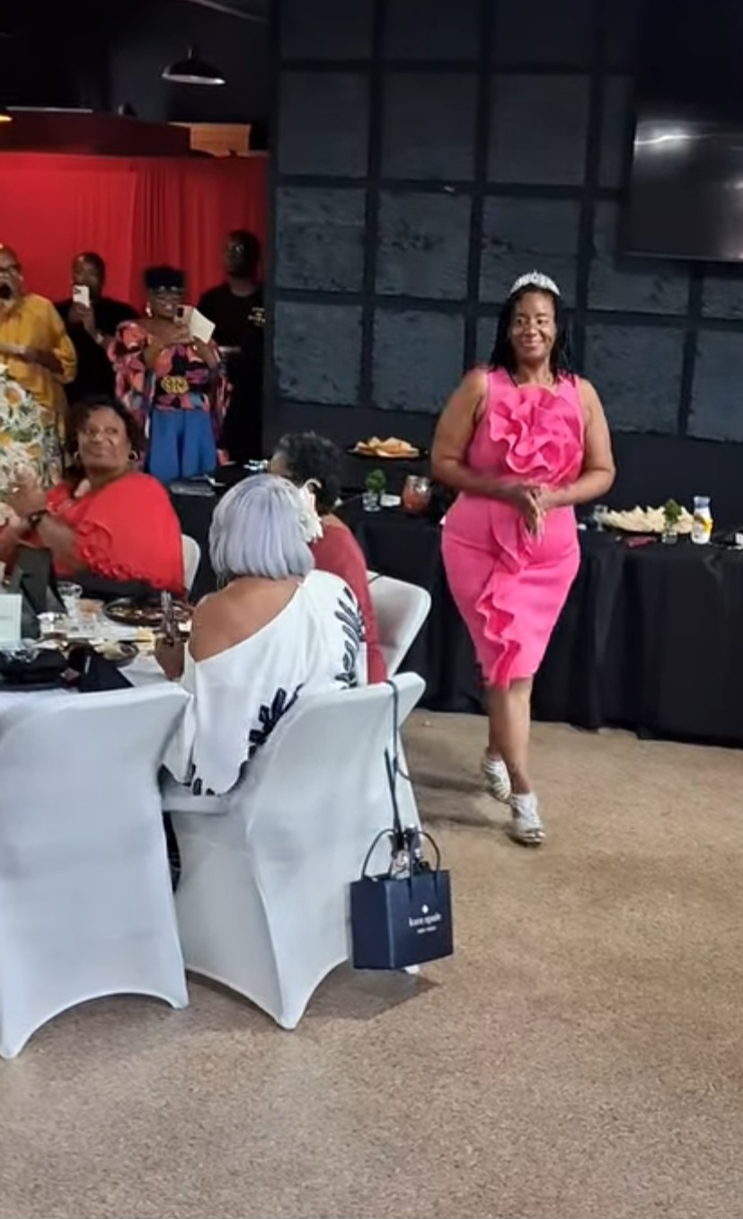 Woman in pink dress walking past seated guests at a social event, with tables and a red curtain in background.