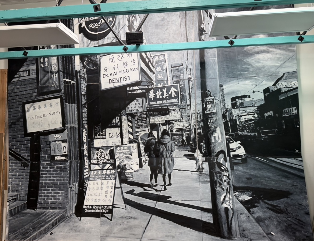 Black-and-white street scene with storefront signs, pedestrians, parked cars, and overhead wires.