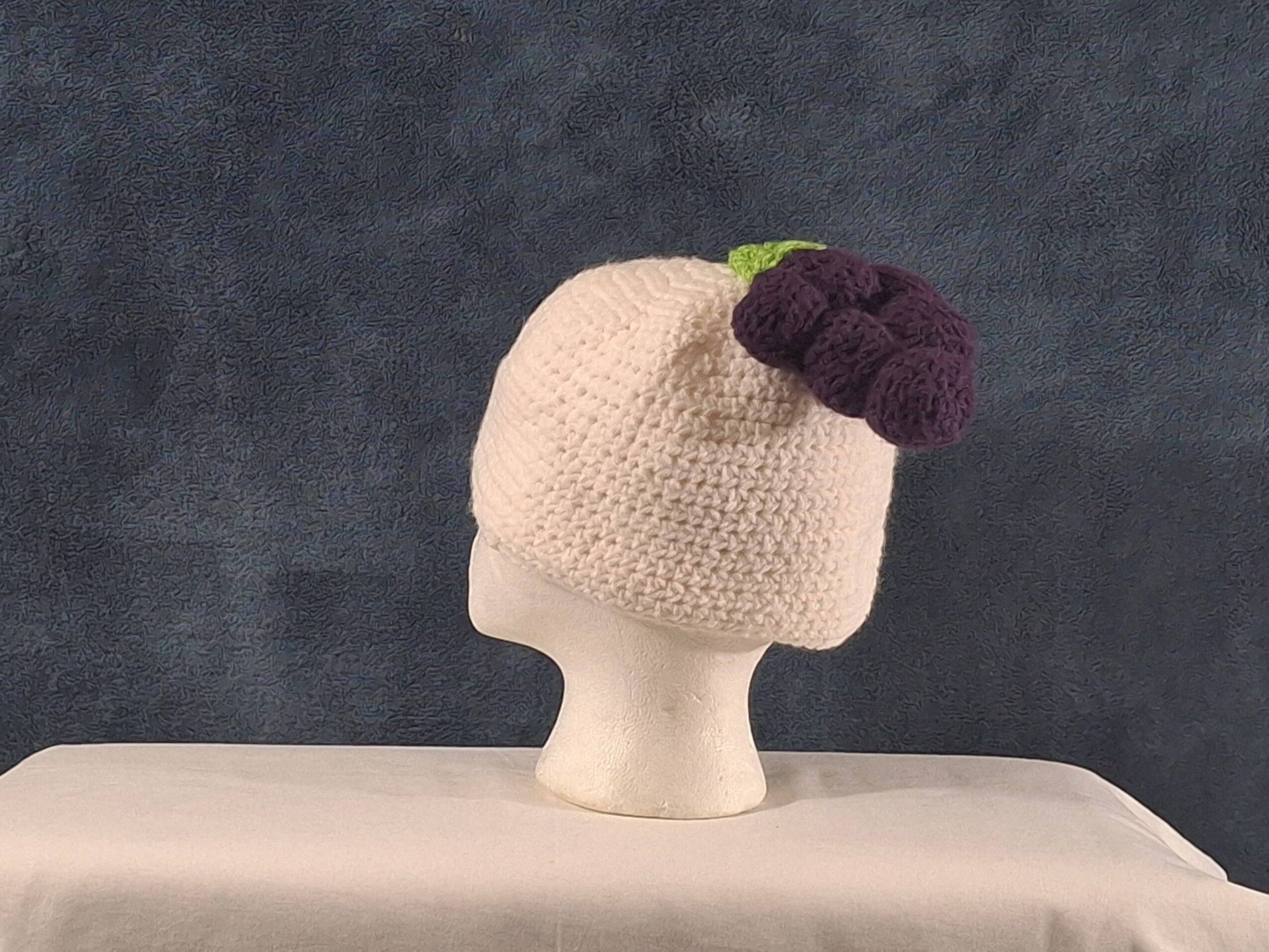 A crocheted beige hat with a purple berry decoration sits on a mannequin head against a dark background.