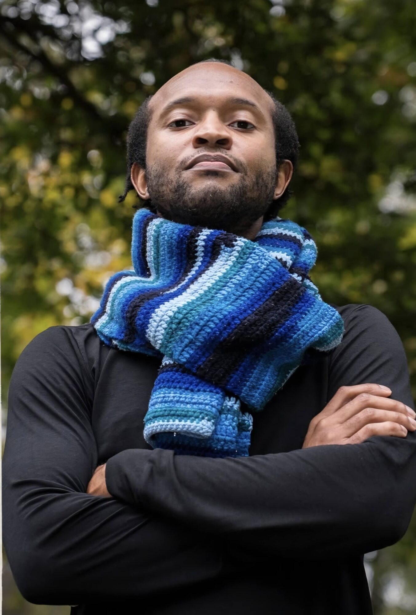 Person wearing a blue striped scarf and black top, standing with arms crossed beneath leafy trees.