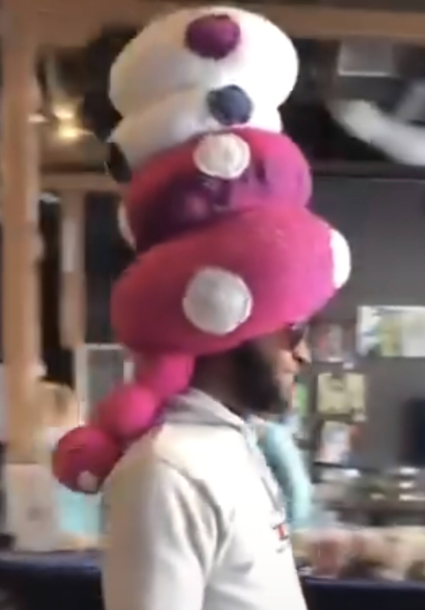 A person in profile wearing a tall stack of colorful plush toys on their head indoors.