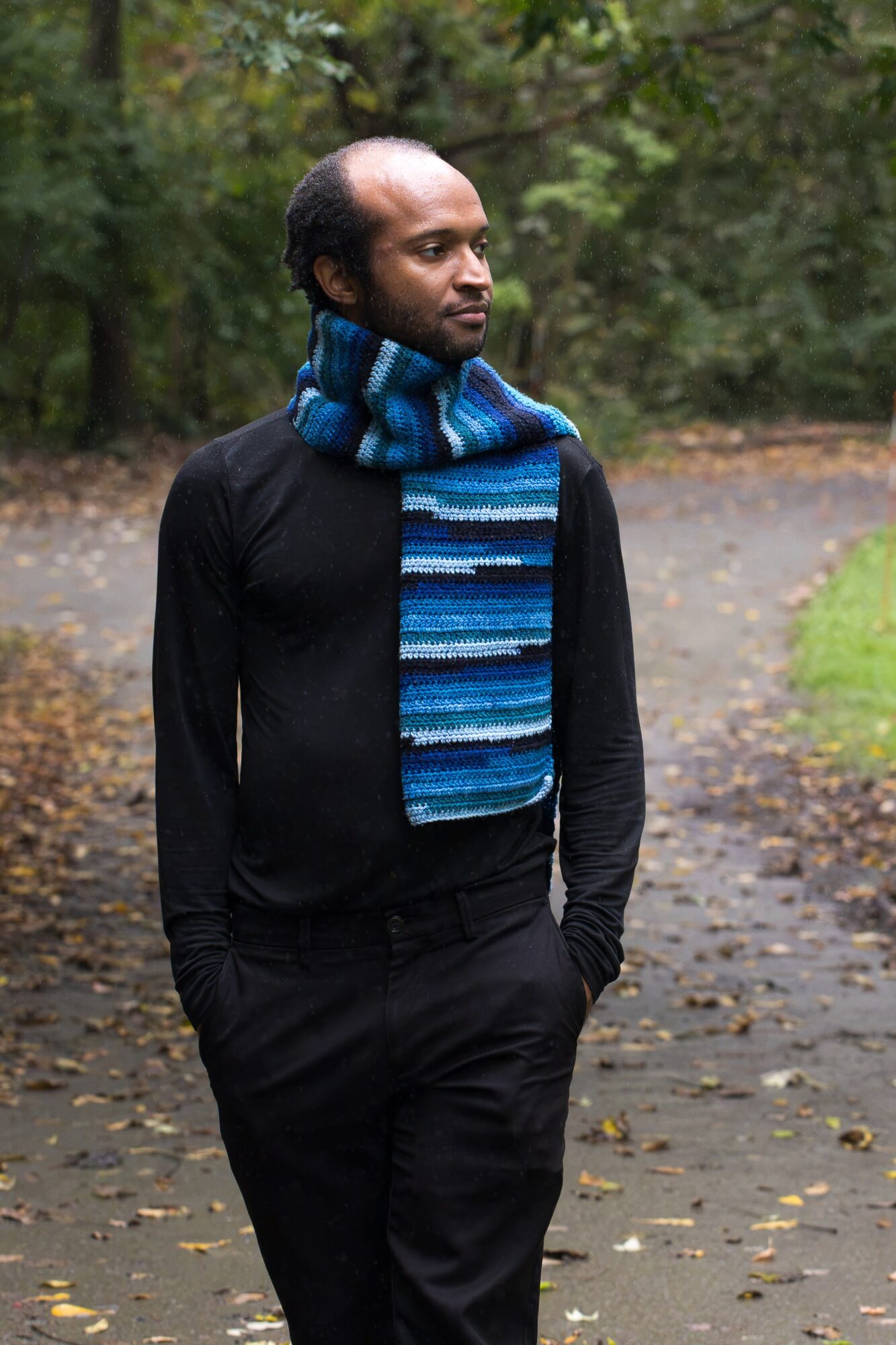A person wearing a blue striped scarf stands on a leaf-strewn path in a wooded area.
