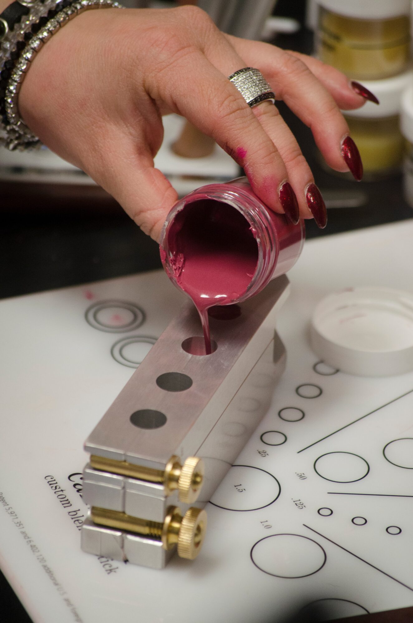 Hand pours pink liquid from a small jar onto a metal tool on a patterned surface.