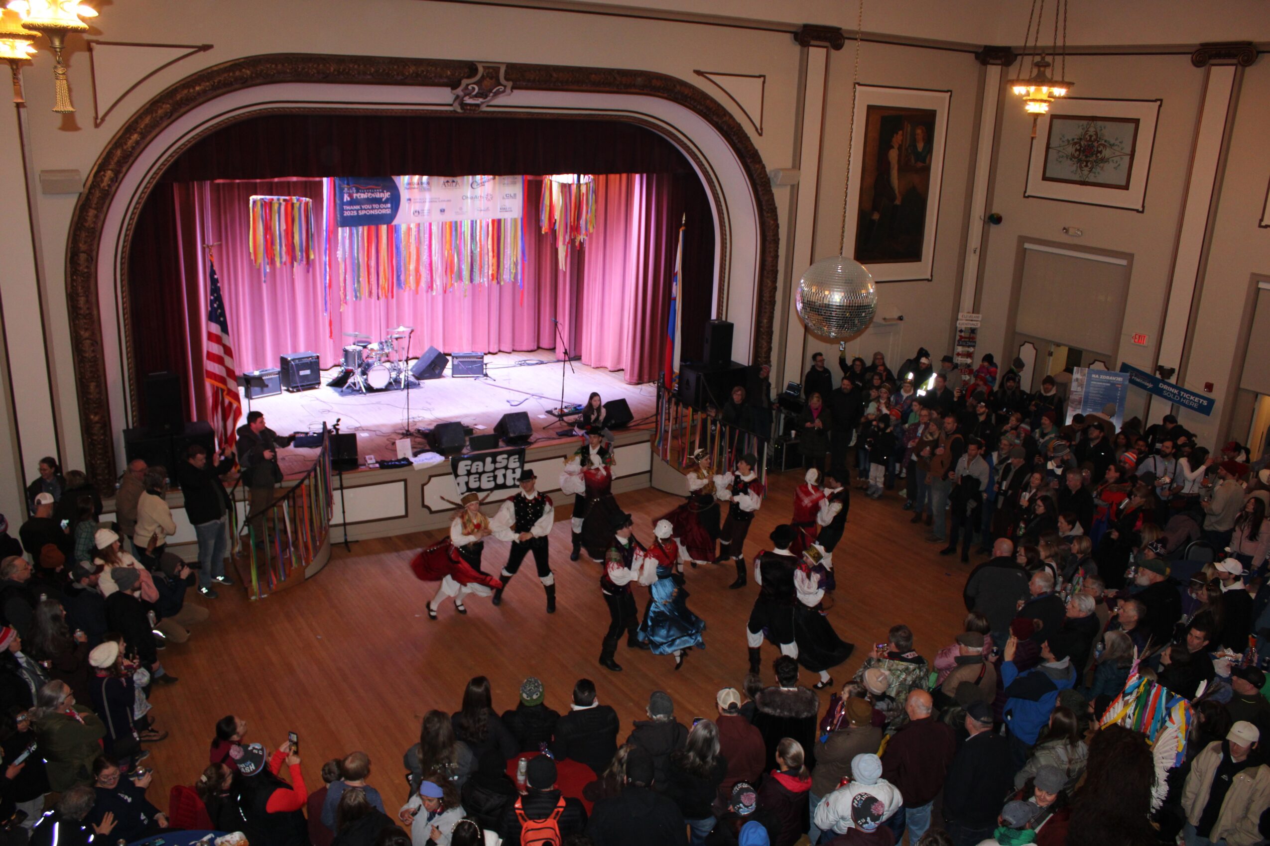 A crowded hall surrounds a stage with pink curtains as performers dance on the floor.