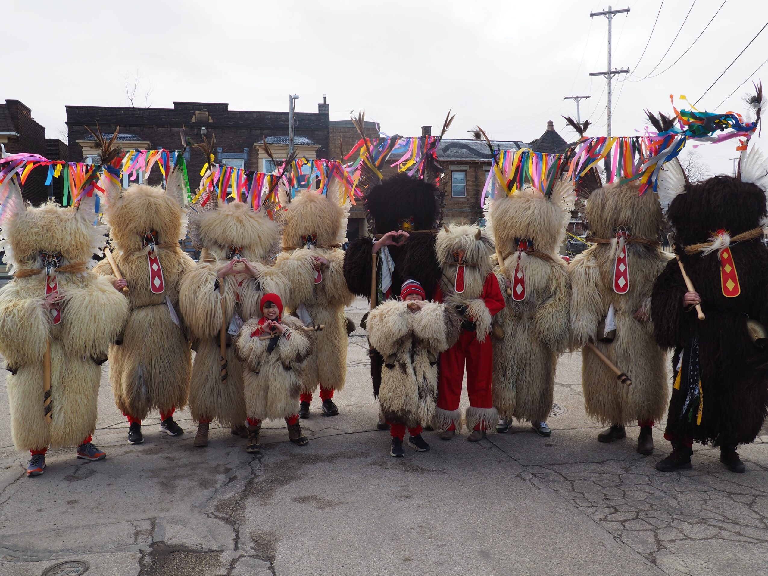 A group of people in shaggy costumes and masks stands beneath colorful streamers on a street.