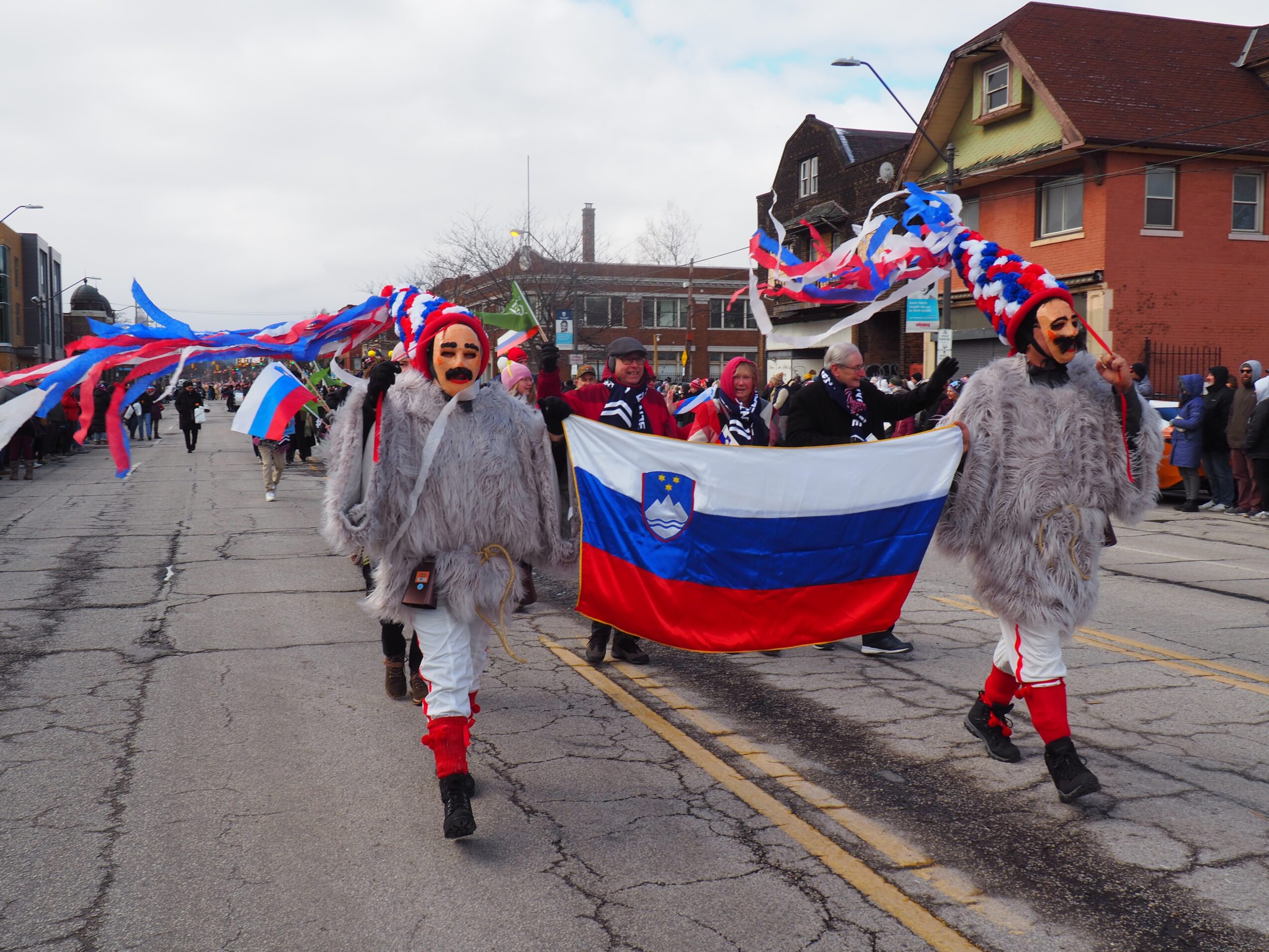 Two costumed participants lead a parade carrying a Slovenian flag, with colorful streamers and spectators along the street.
