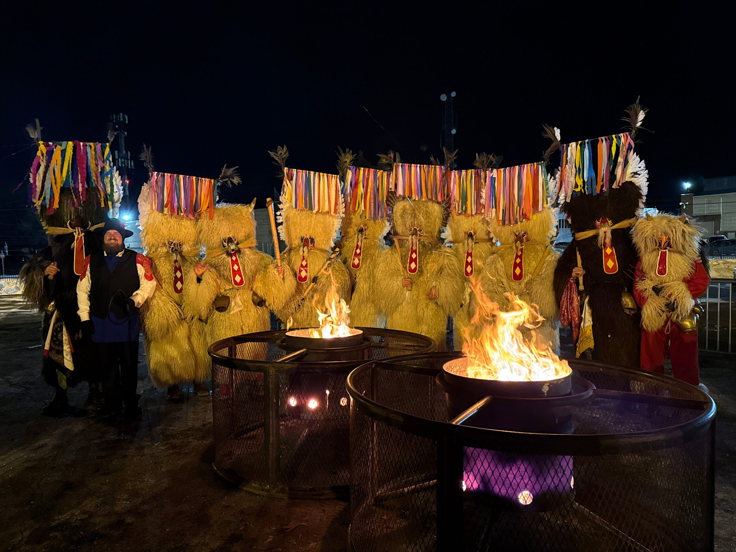 Two fire pits burn before straw-covered figures decorated with colorful ribbons and ornaments at night.