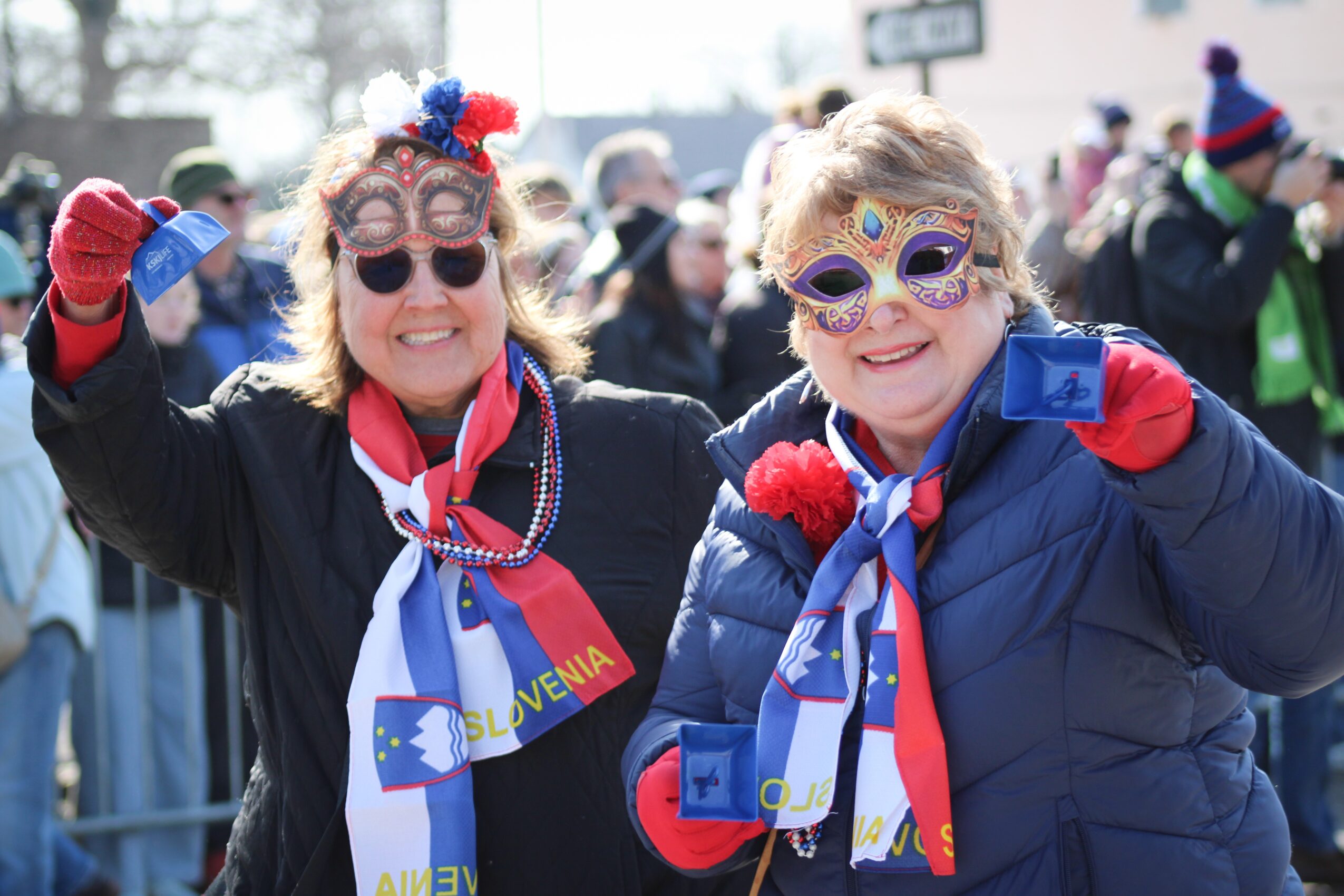 Two people wearing colorful glasses, scarves, and winter jackets wave at an outdoor event.