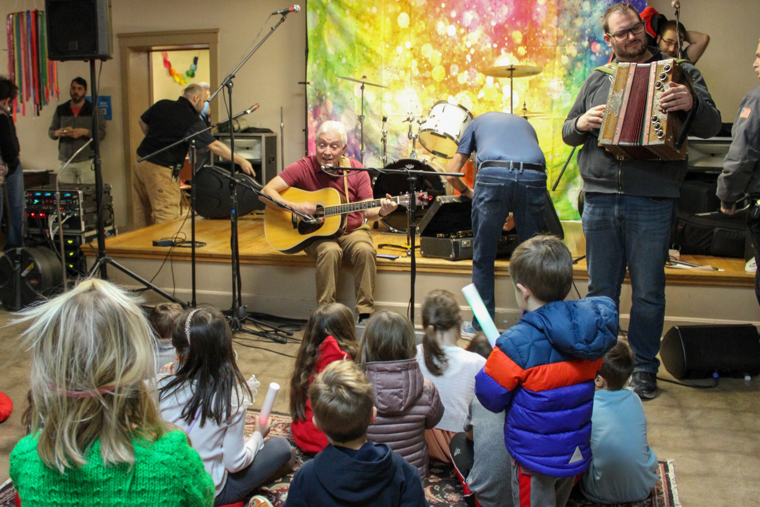 Musicians perform on a small stage while seated children watch from the floor.