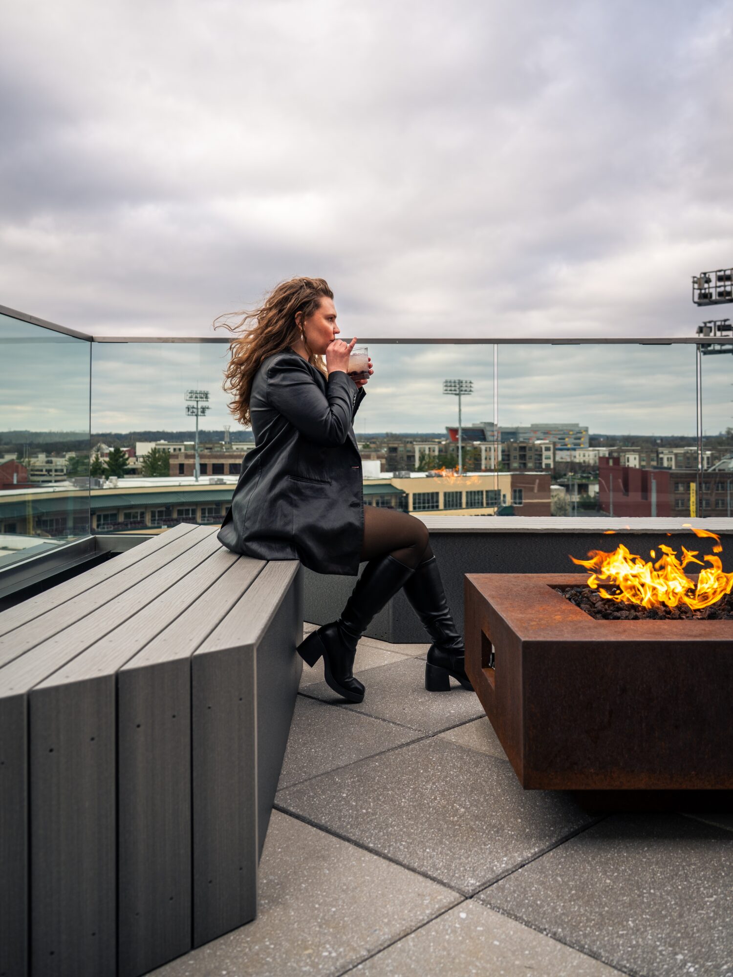 A person sits beside a lit fire pit on a rooftop terrace under a cloudy sky.