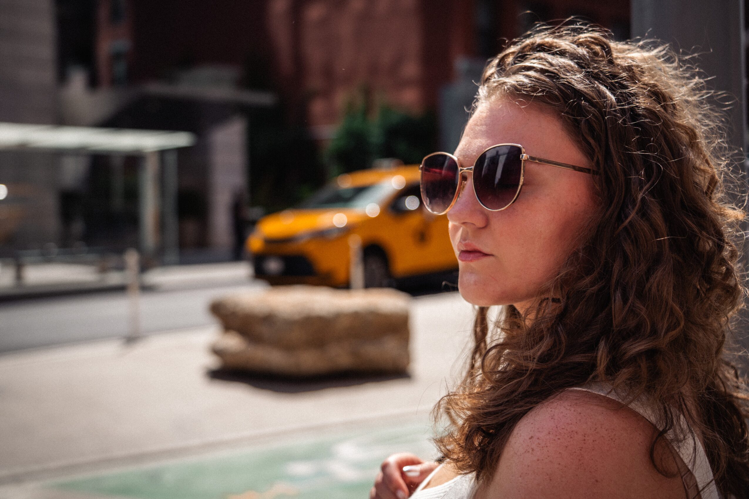 Person wearing sunglasses stands beside a city street, with a yellow vehicle and planter in the background.
