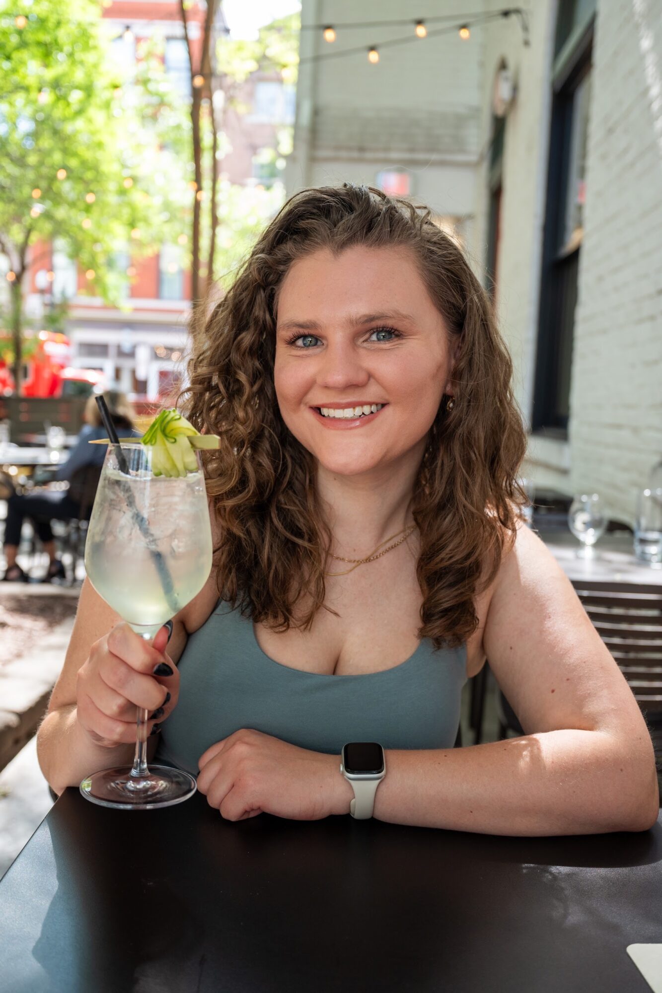 Person seated at an outdoor restaurant table, holding a glass of white wine with a straw.