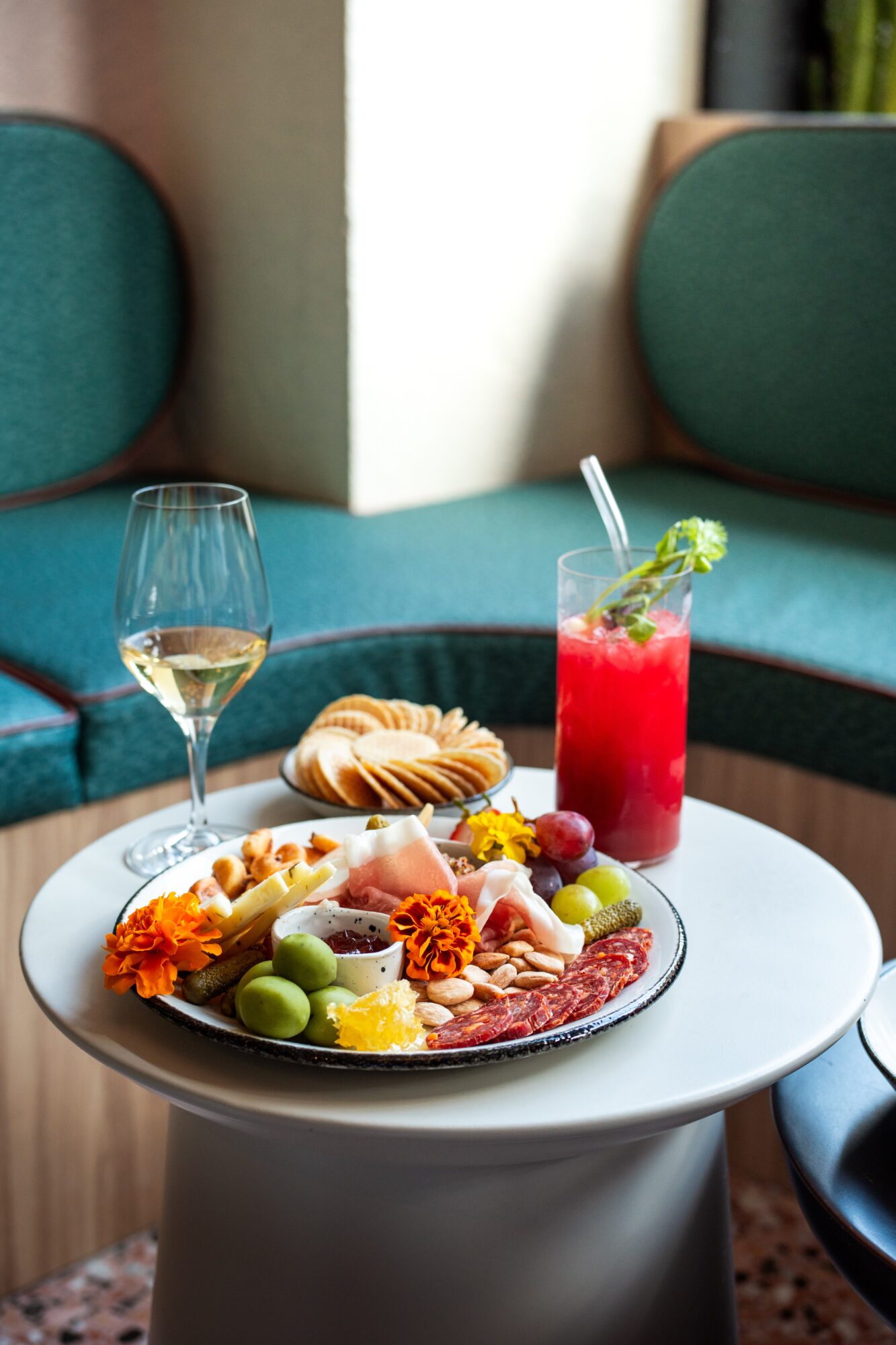 A platter of appetizers with a wine glass and red drink on a round table between teal chairs.