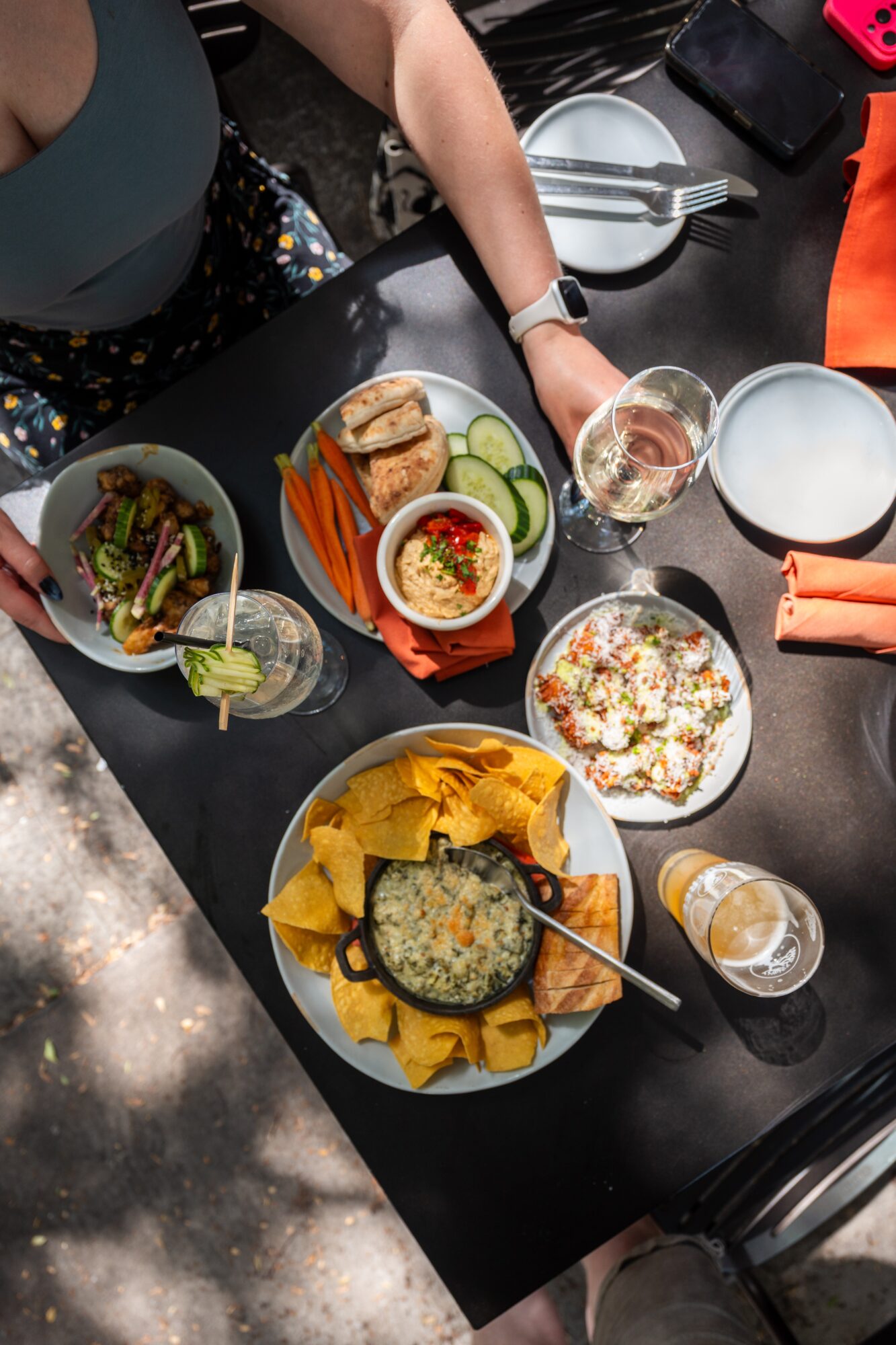 Overhead view of a table with plates of food, drinks, and people’s arms reaching toward the dishes.