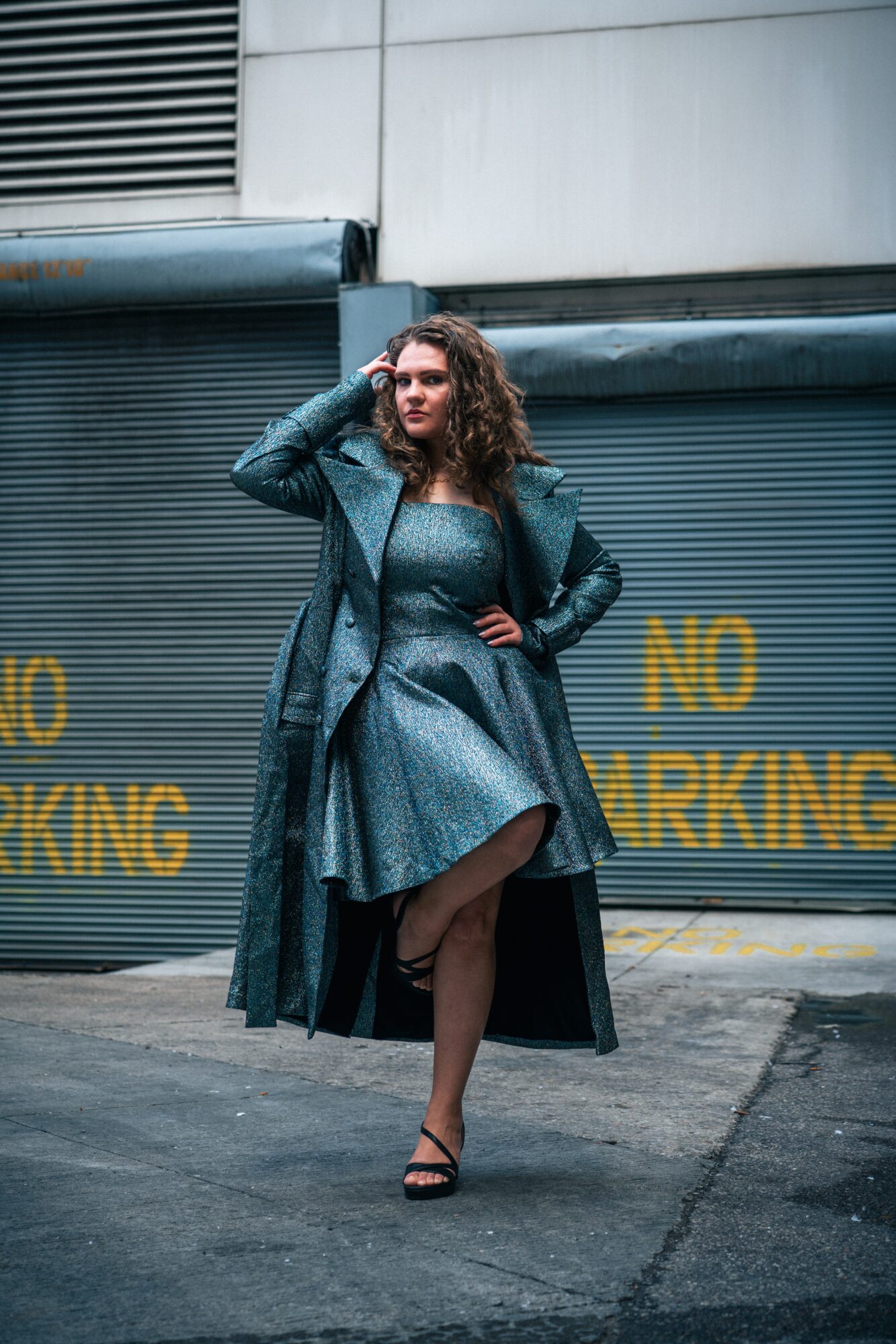 Person in a long teal coat poses in front of closed metal shutters with yellow lettering.