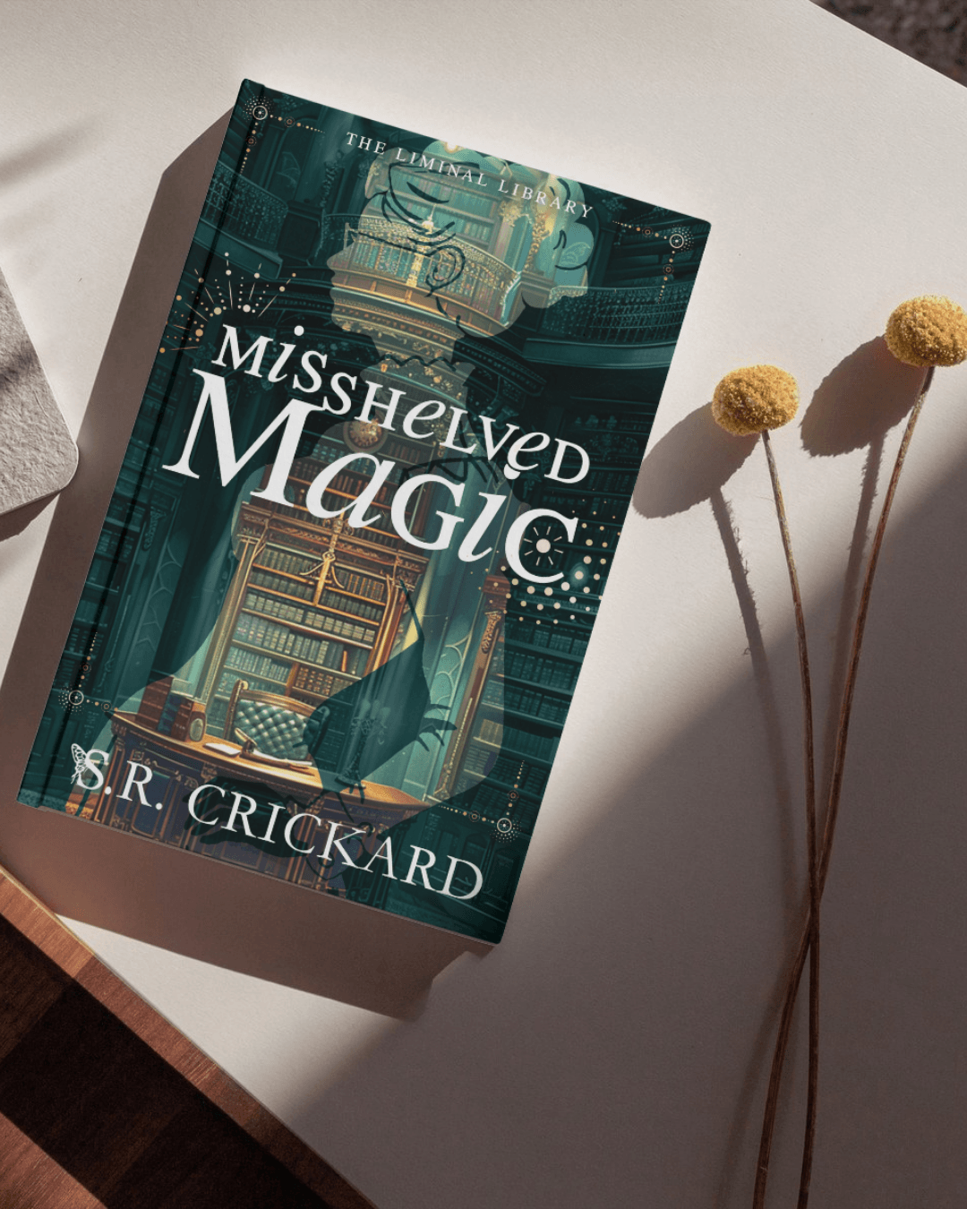 A teal hardcover book titled “Misshelved Magic” lies beside three dried yellow flowers.