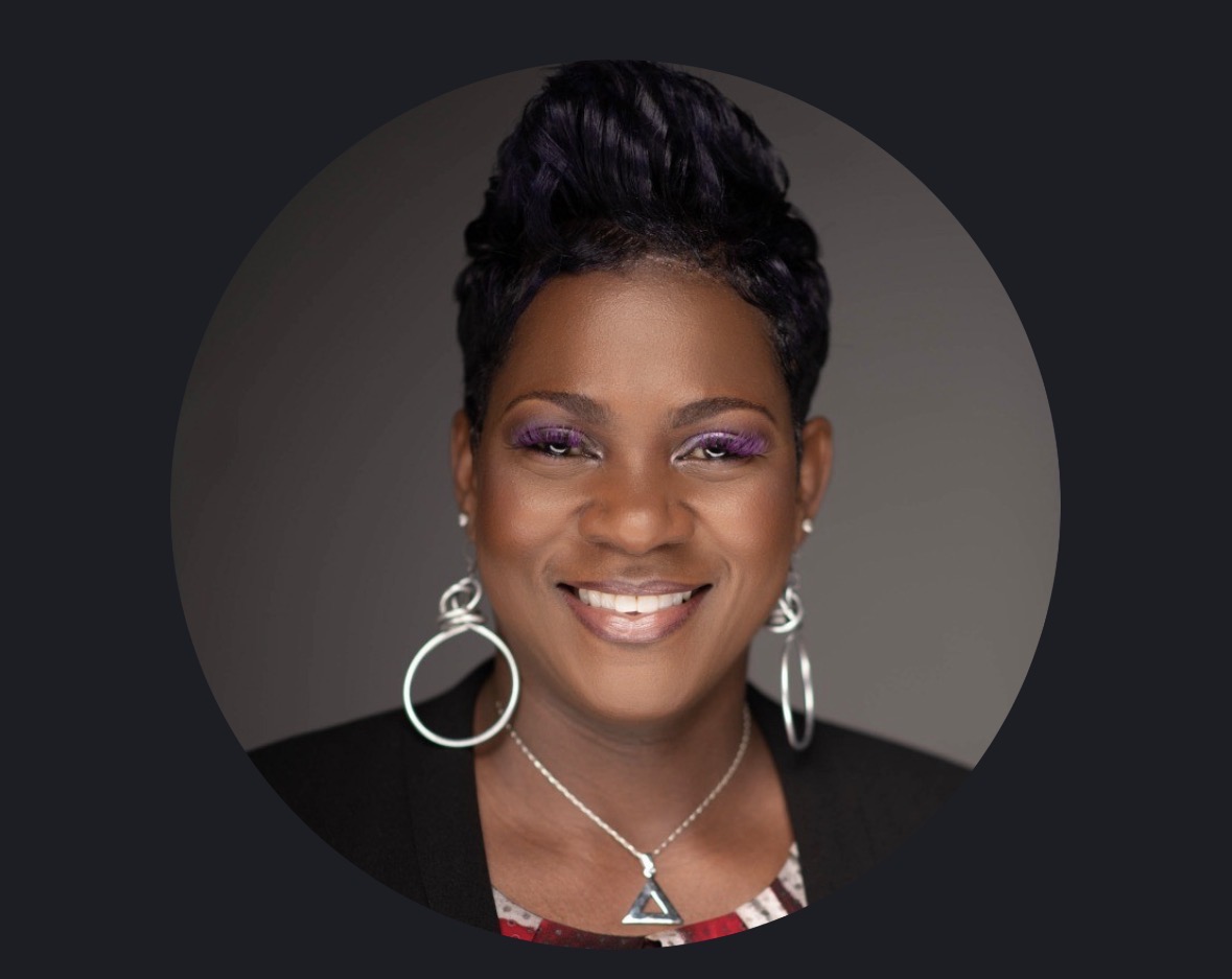 Portrait of a smiling person with an updo hairstyle, hoop earrings, and a necklace against a dark background.