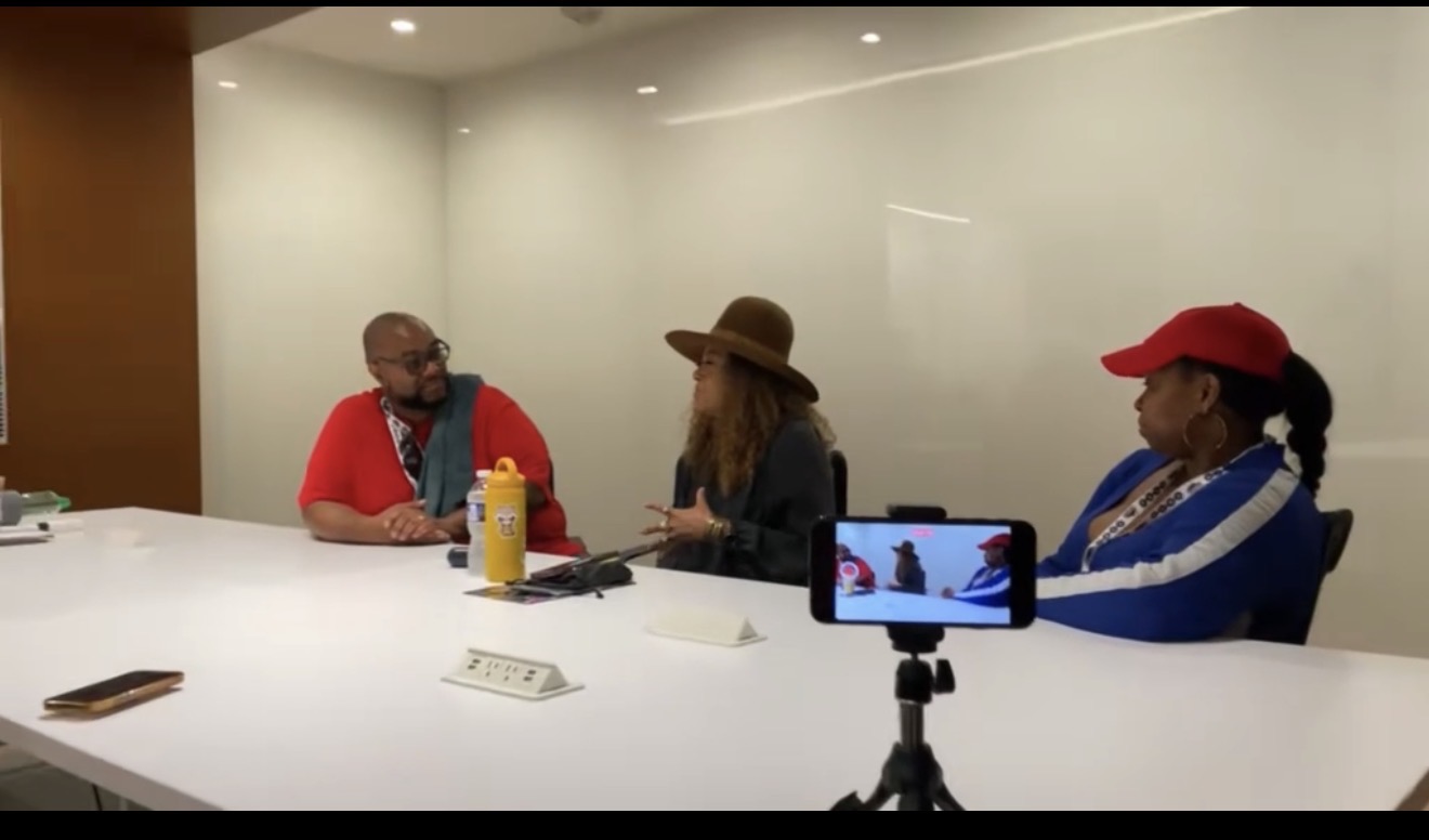 Three people sit at a white conference table in a room with plain walls, one person is recording with a phone on a tripod.