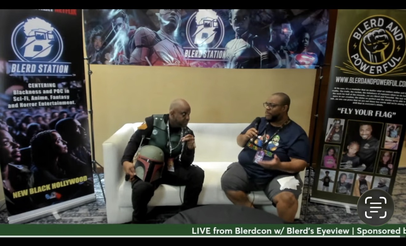 Two men sit on a white couch, talking, with banners and posters behind them at an event or interview setting.