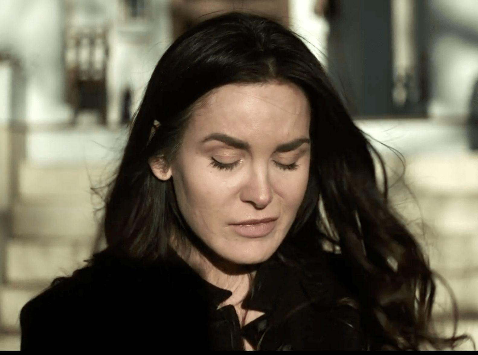 A person with long dark hair and closed eyes, wearing a black coat outdoors.