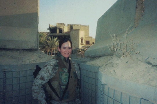 A person wearing camouflage and a helmet stands in a sandbagged trench, with buildings in the background.