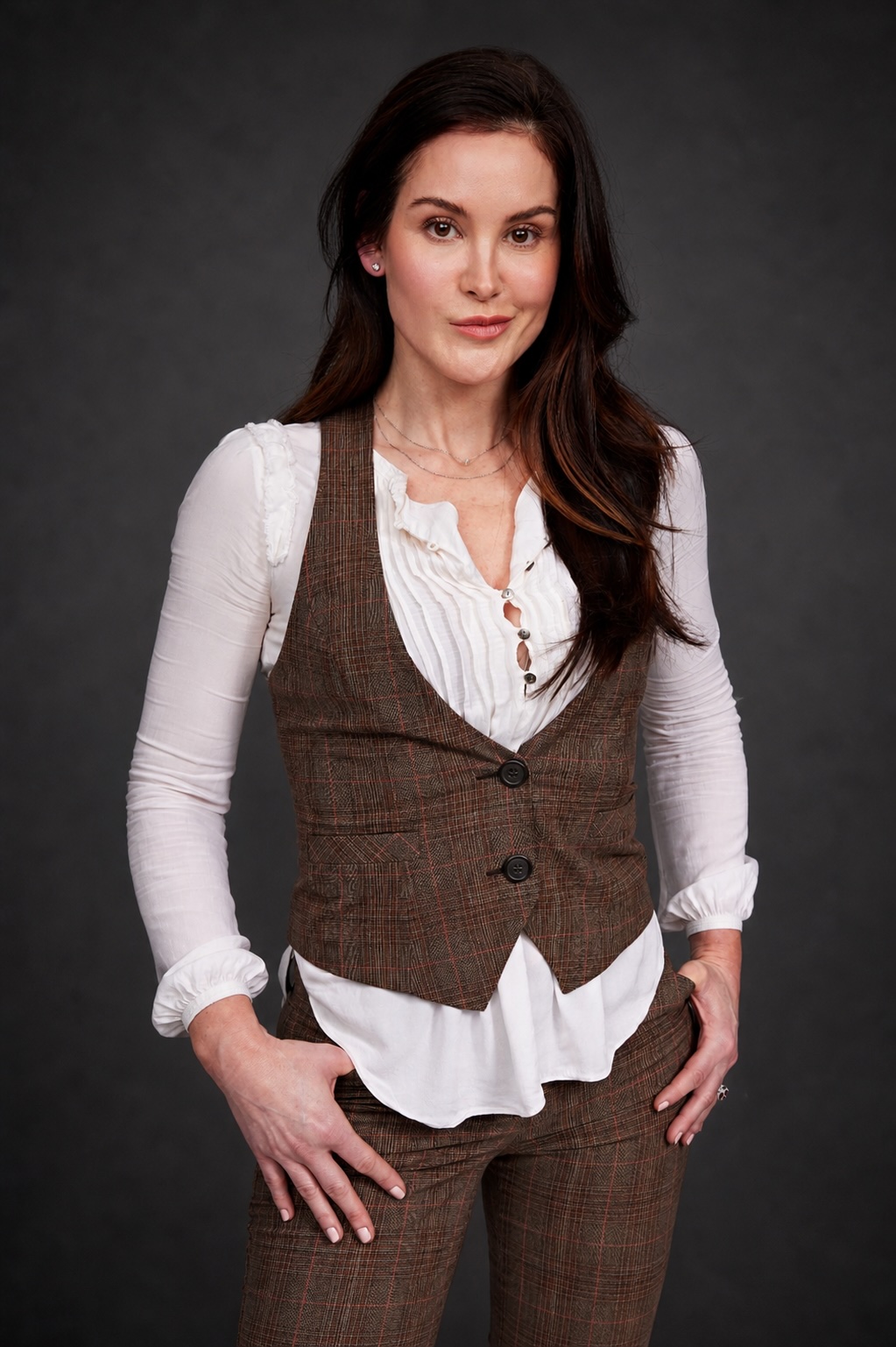 A person with long dark hair wearing a white blouse and brown vest and trousers against a dark background.