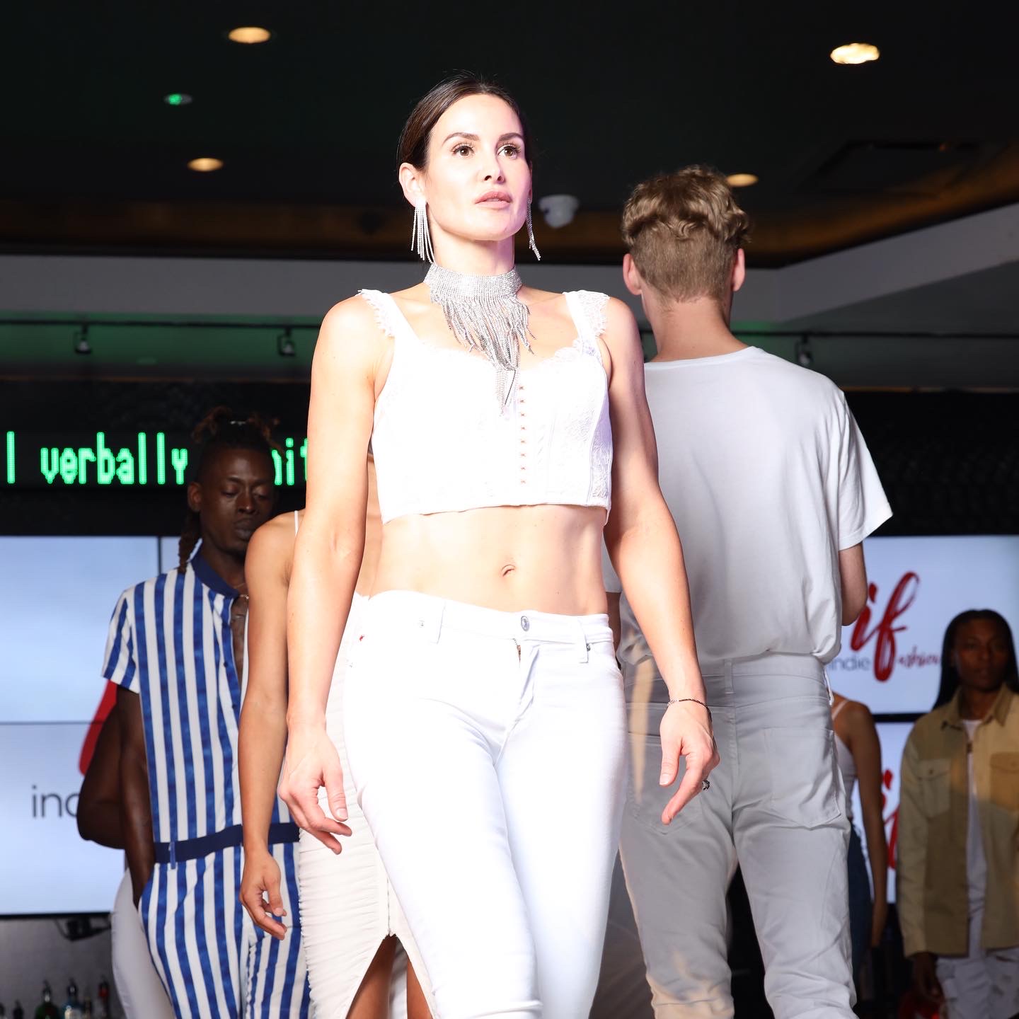 A woman in white walks on a runway as people stand behind her.