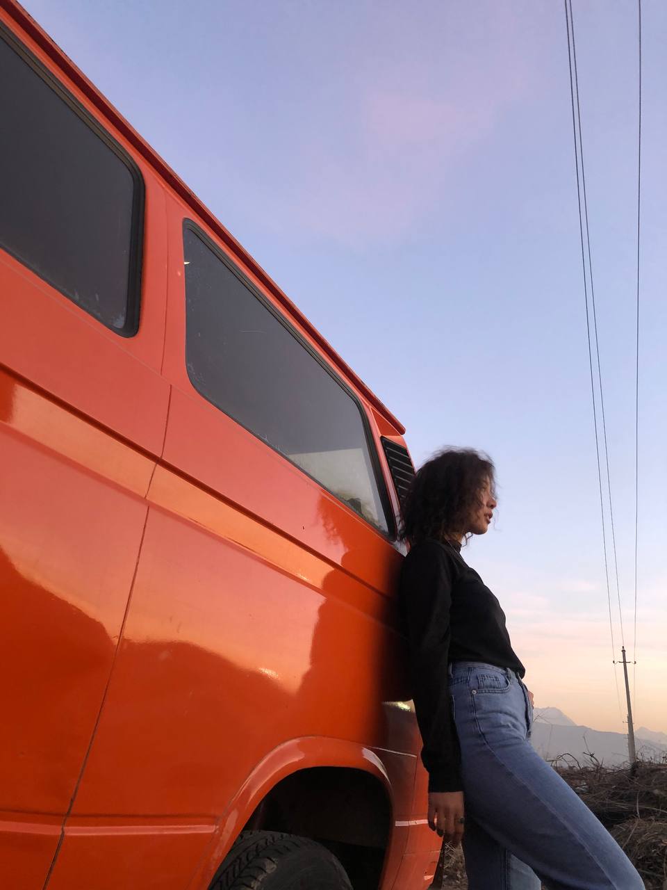 Person leaning against an orange van beside a roadside at sunset.