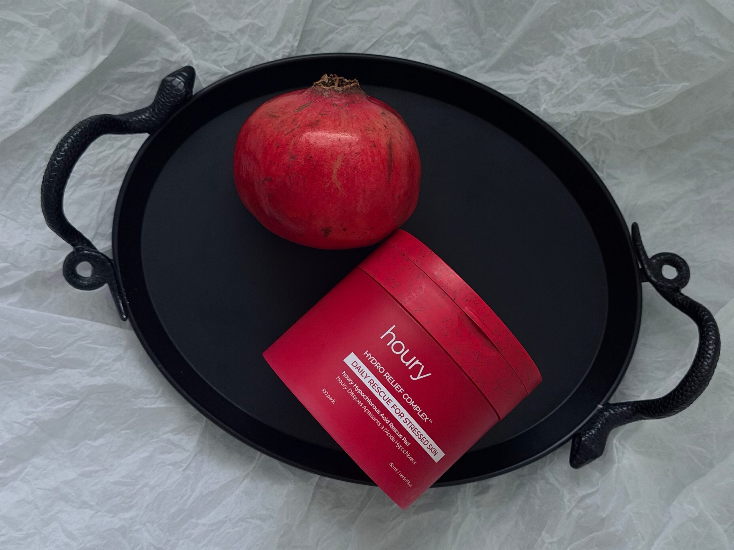 A black pan holds a red pomegranate and a red rectangular box on a light fabric surface.