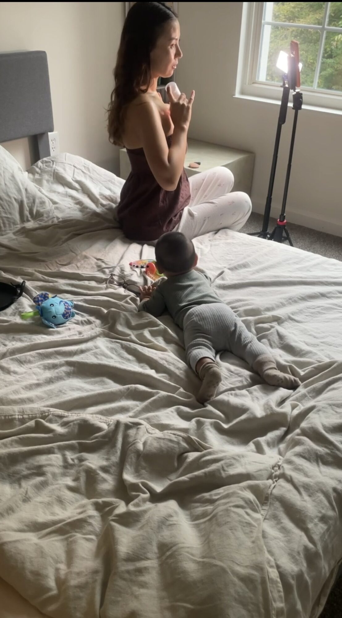 A seated person holds a phone near a window while a baby lies on the bed.