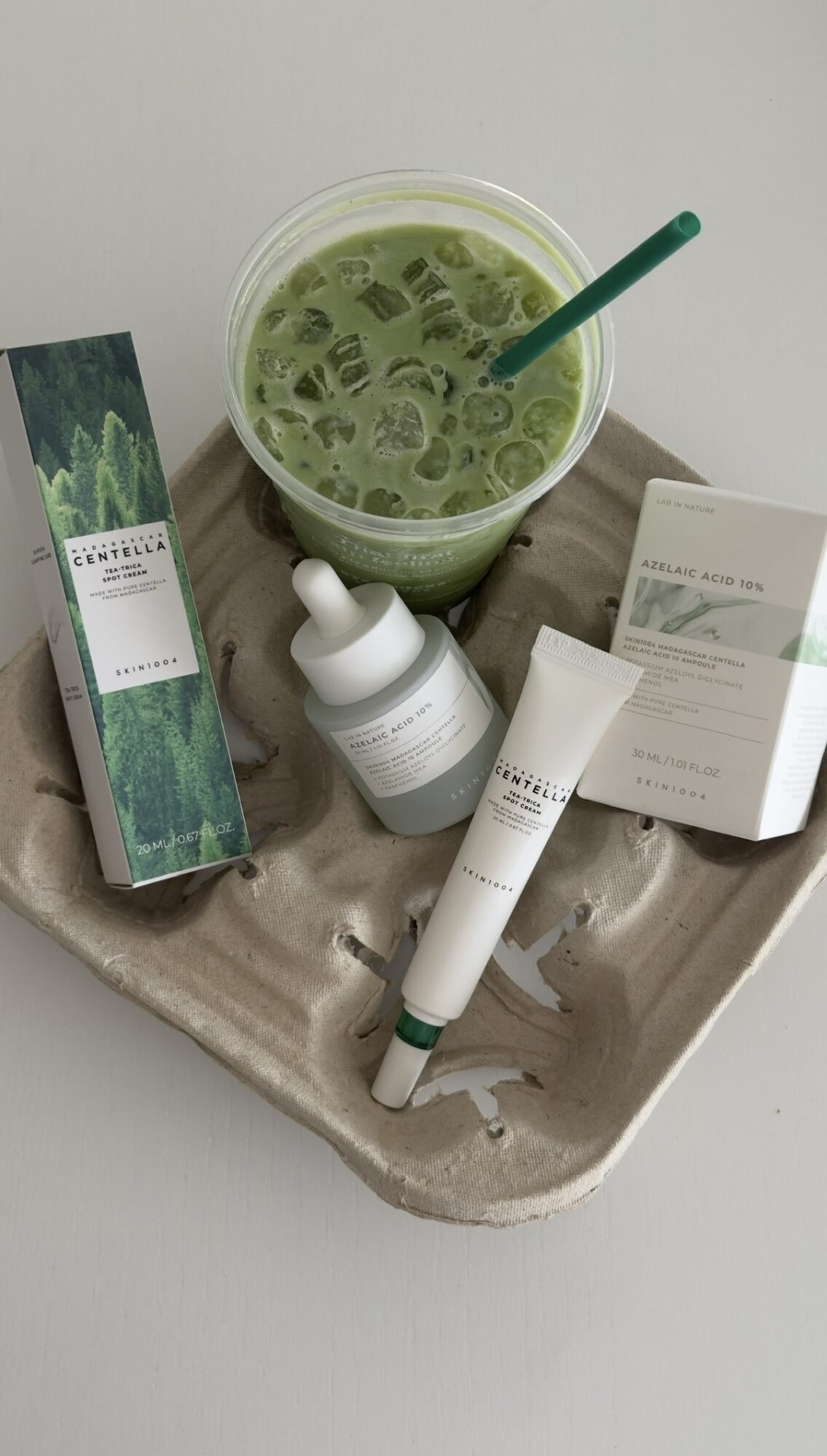 Green face mask in a bowl with a spatula, surrounded by skincare product boxes and tubes on a tray.