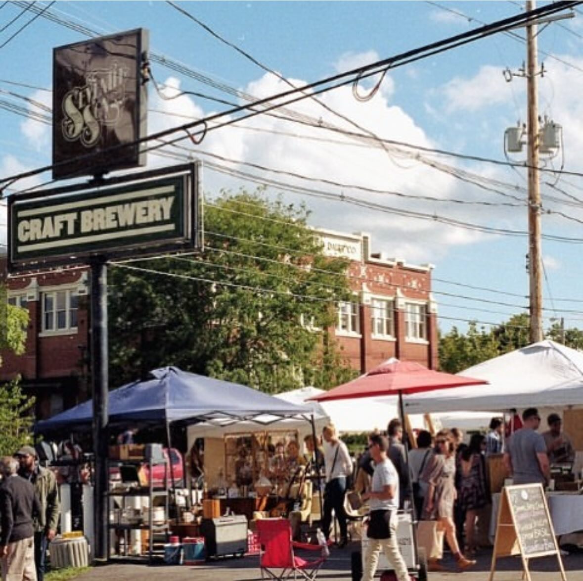 Outdoor craft brewery market with people, vendor tents, a brick building, and overhead utility wires.