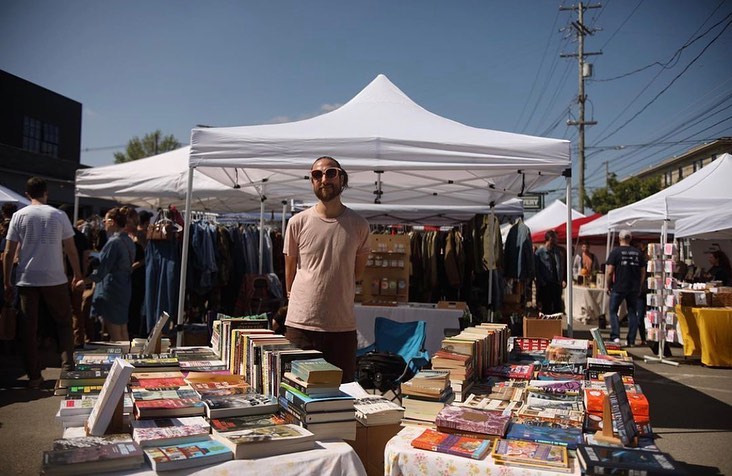 A person stands behind tables of books beneath a white canopy at an outdoor market.